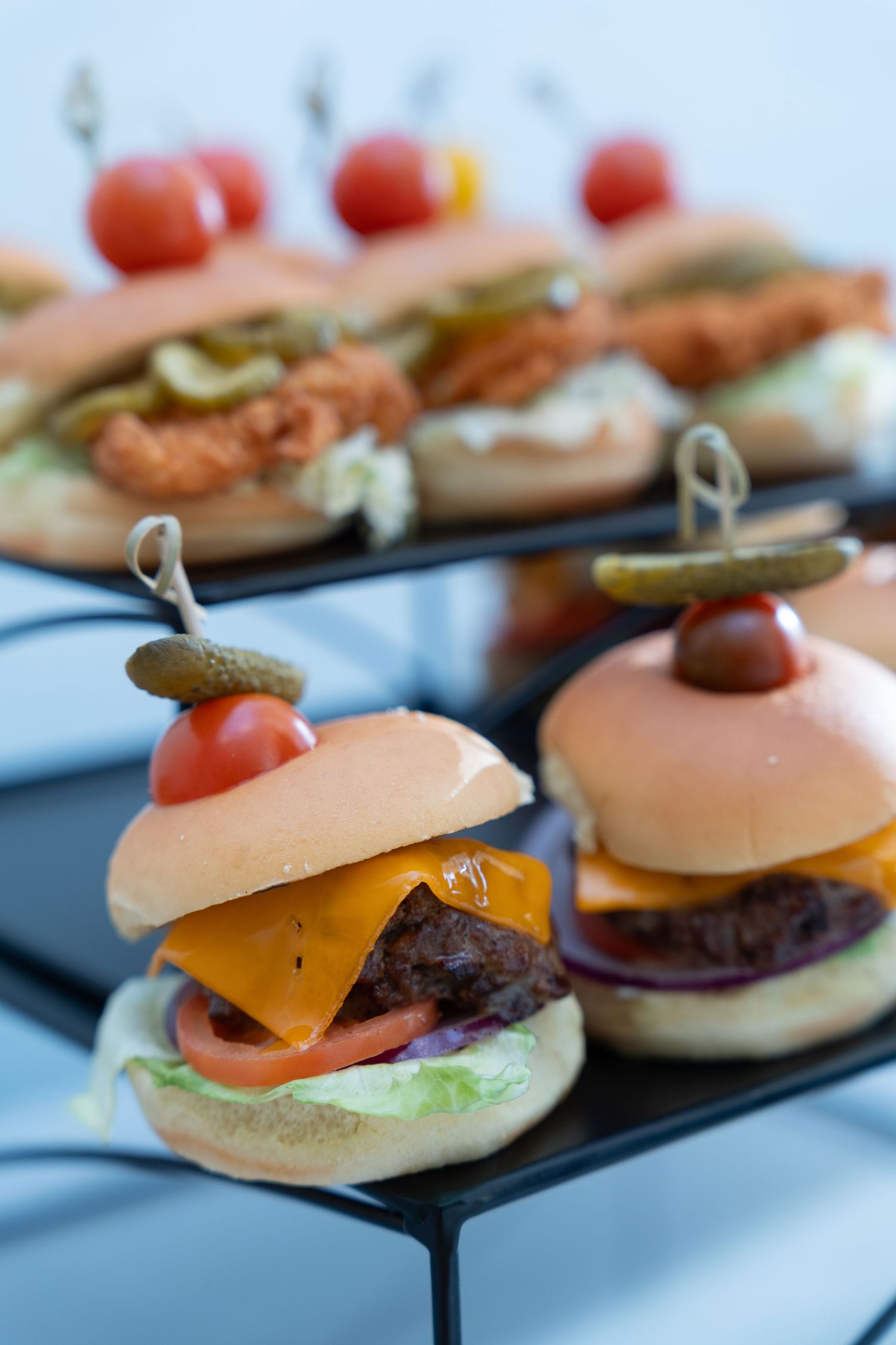 Gourmet mini cheeseburger sliders with cherry tomatoes served on black display stands.