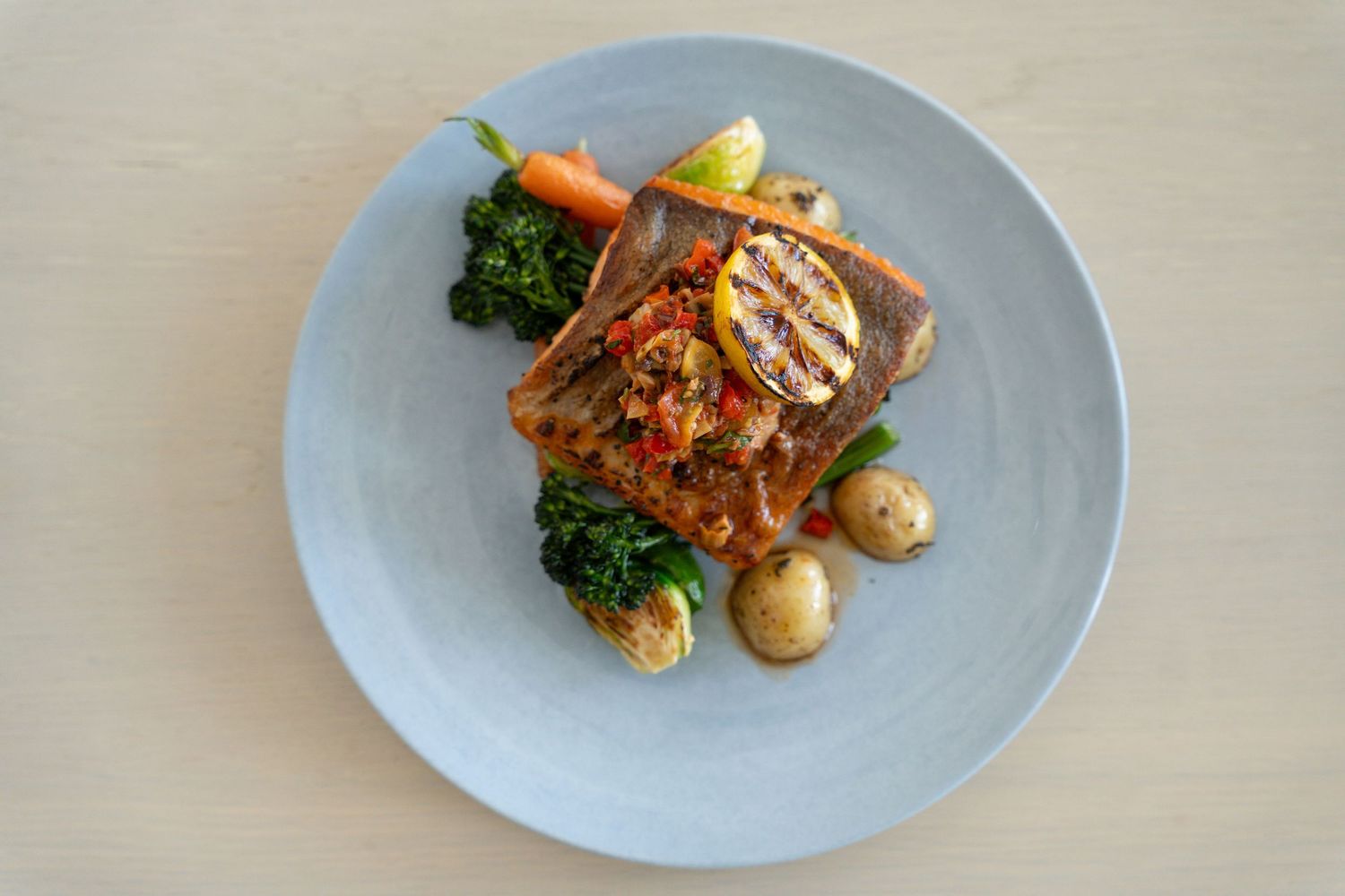 Grilled fish fillet served with roasted potatoes, broccoli, and a vibrant tomato sauce on a blue plate.