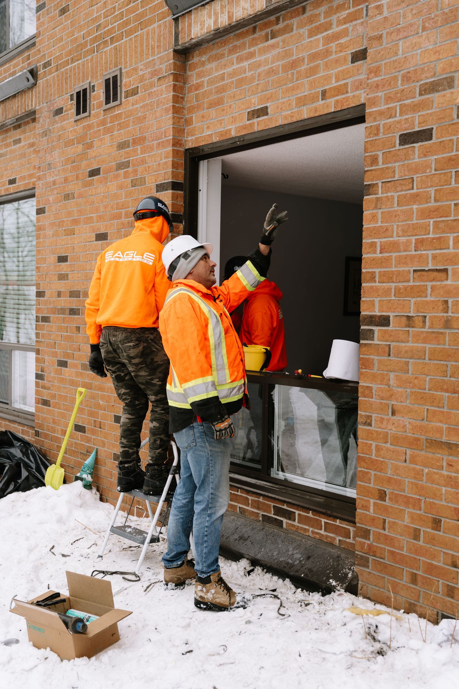 Workers in high-visibility orange jackets install windows on a snowy day at a brick building.