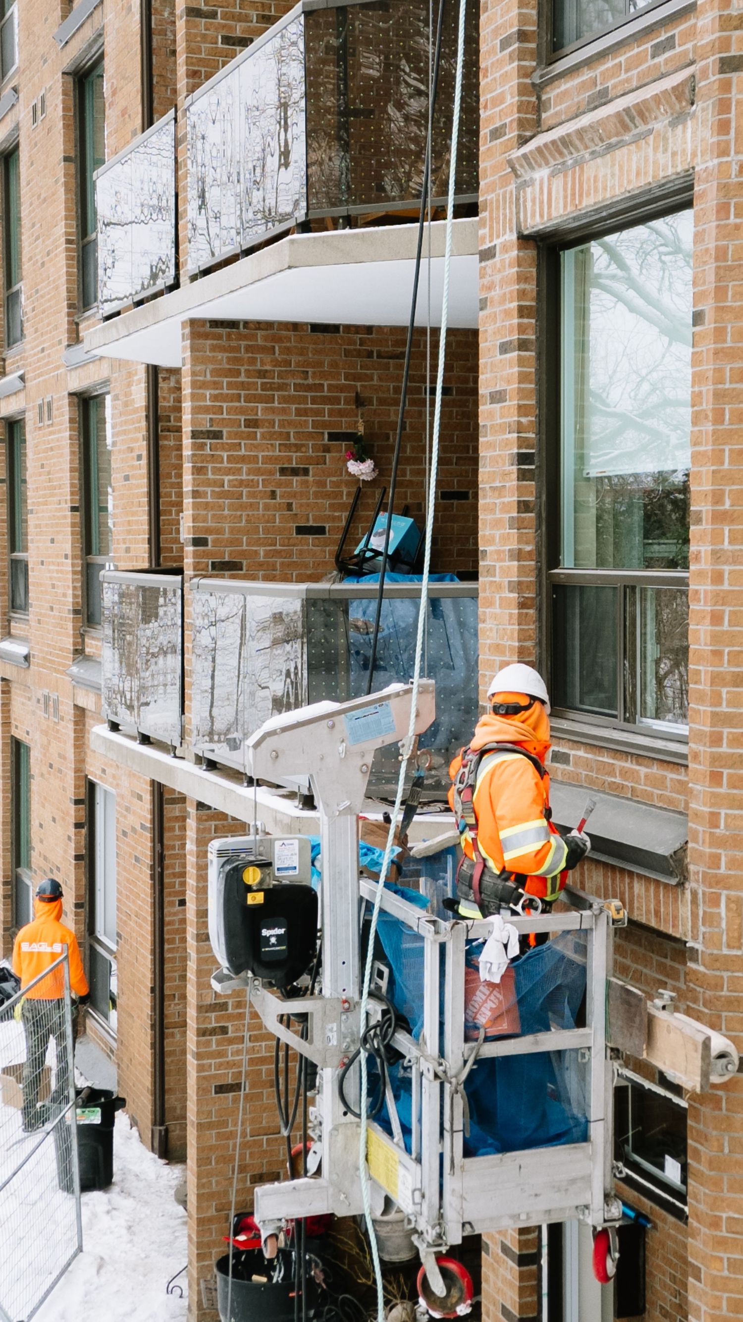Construction workers in orange safety vests operate a suspended platform lift while working on a brick building exterior.