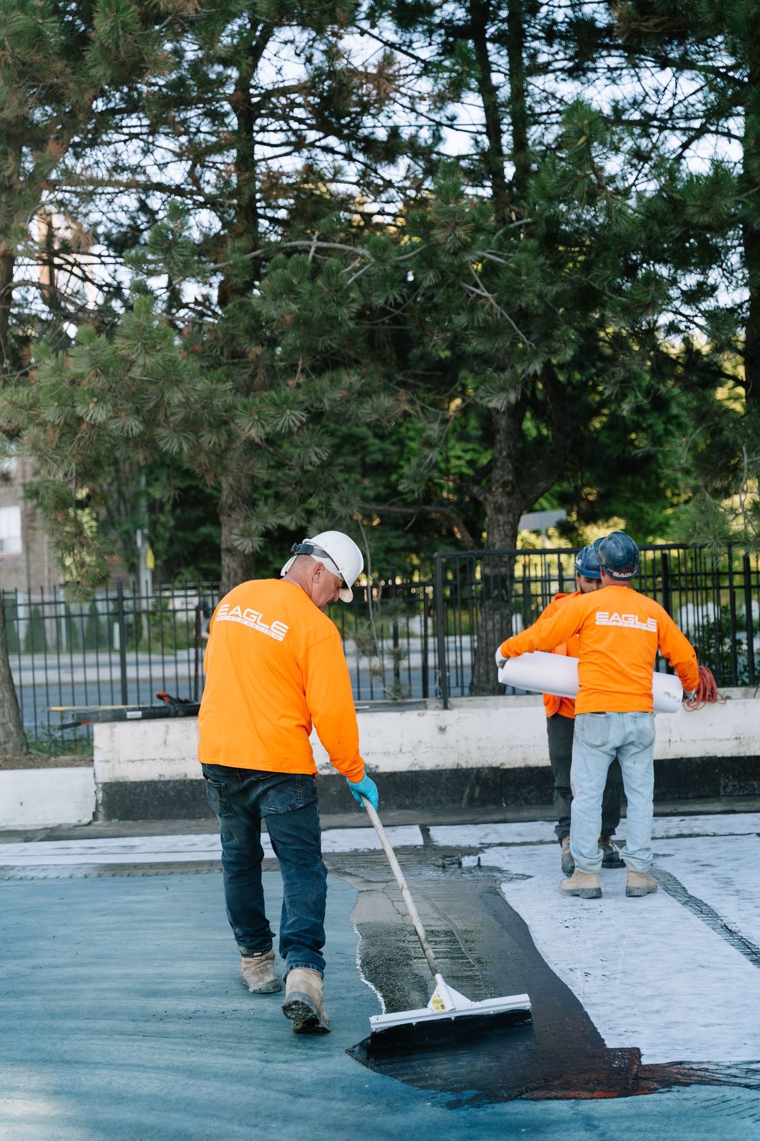 Maintenance crew in bright orange vests applying dark coating to a paved surface.