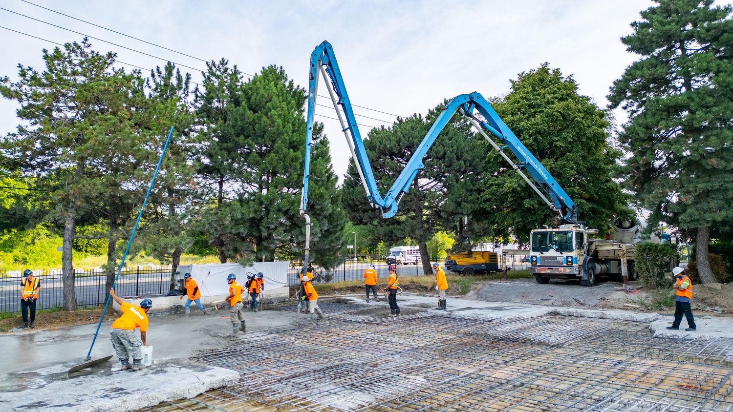 Construction workers and cranes work on a large concrete foundation at an outdoor building site on a sunny day.