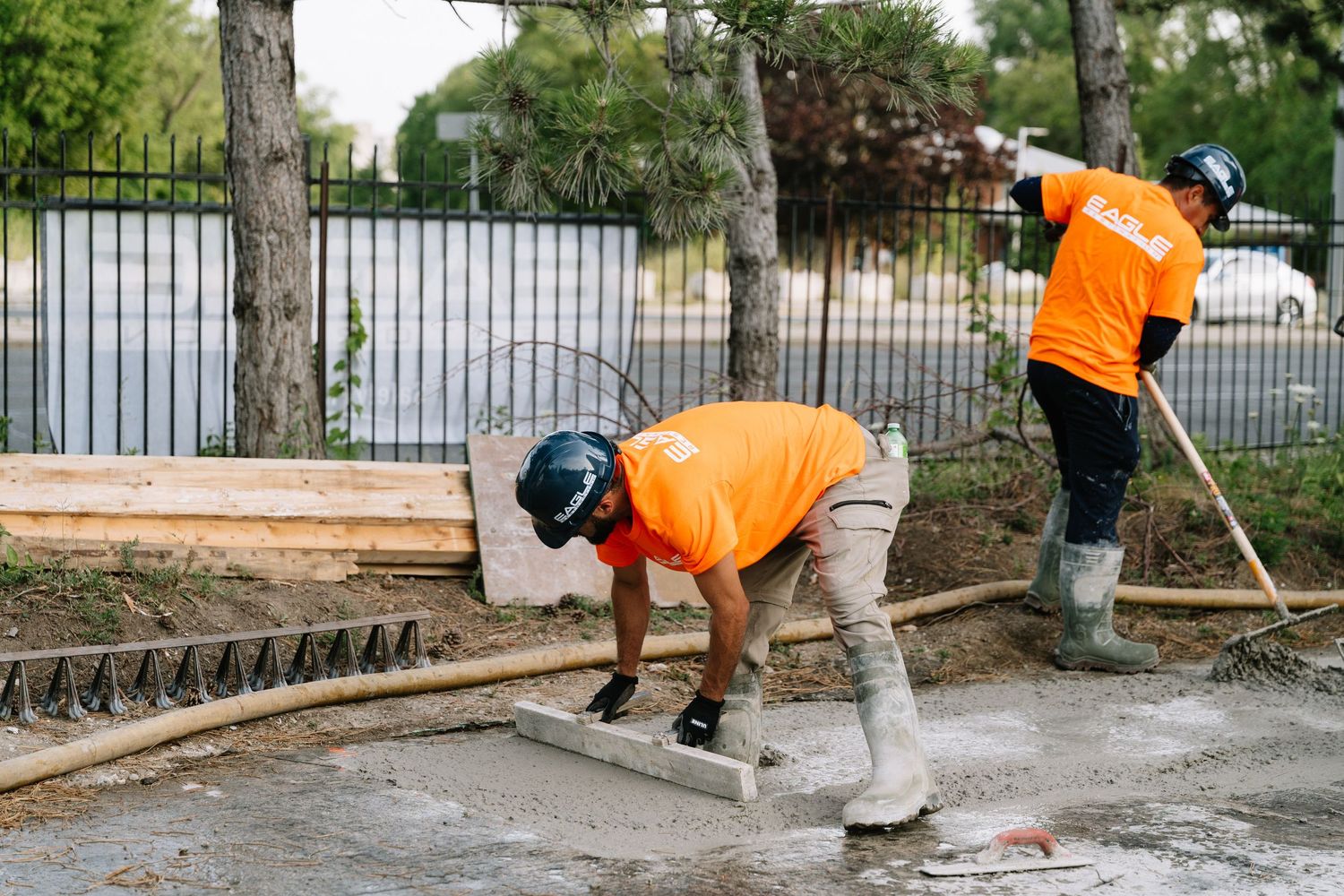 Construction workers in orange vests laying concrete near a metal fence and trees.