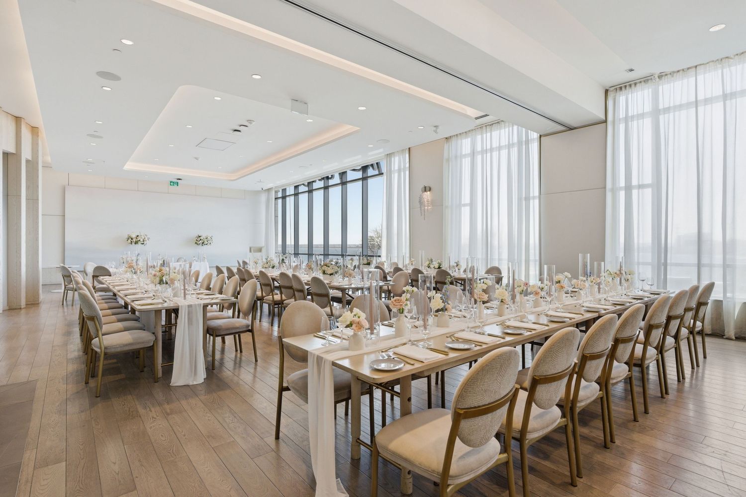 Modern banquet hall with white decor features long dining tables, wooden chairs, and floor-to-ceiling windows overlooking city views.