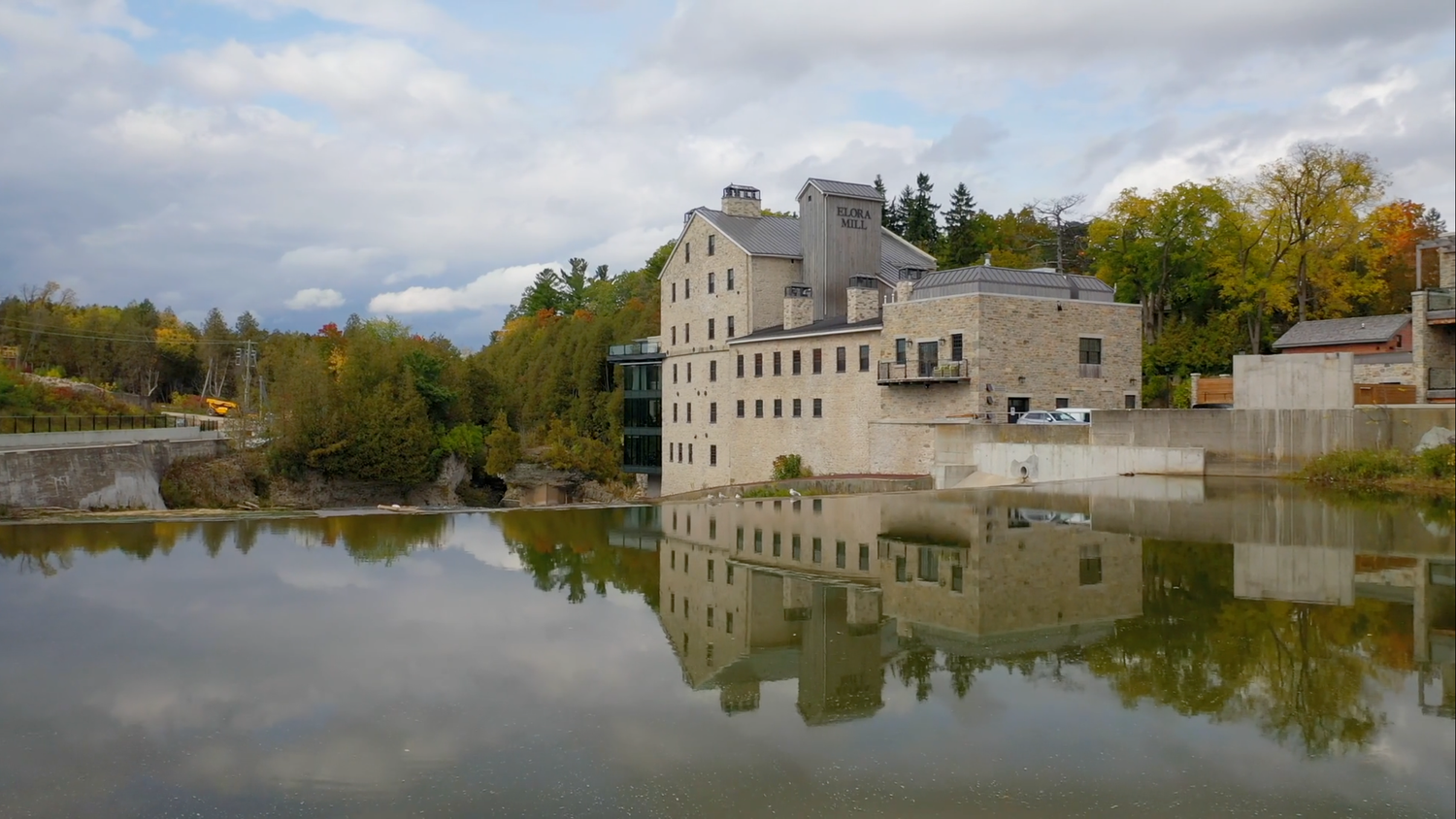 Historic stone mill building reflected in calm water, surrounded by autumn trees under a cloudy sky.