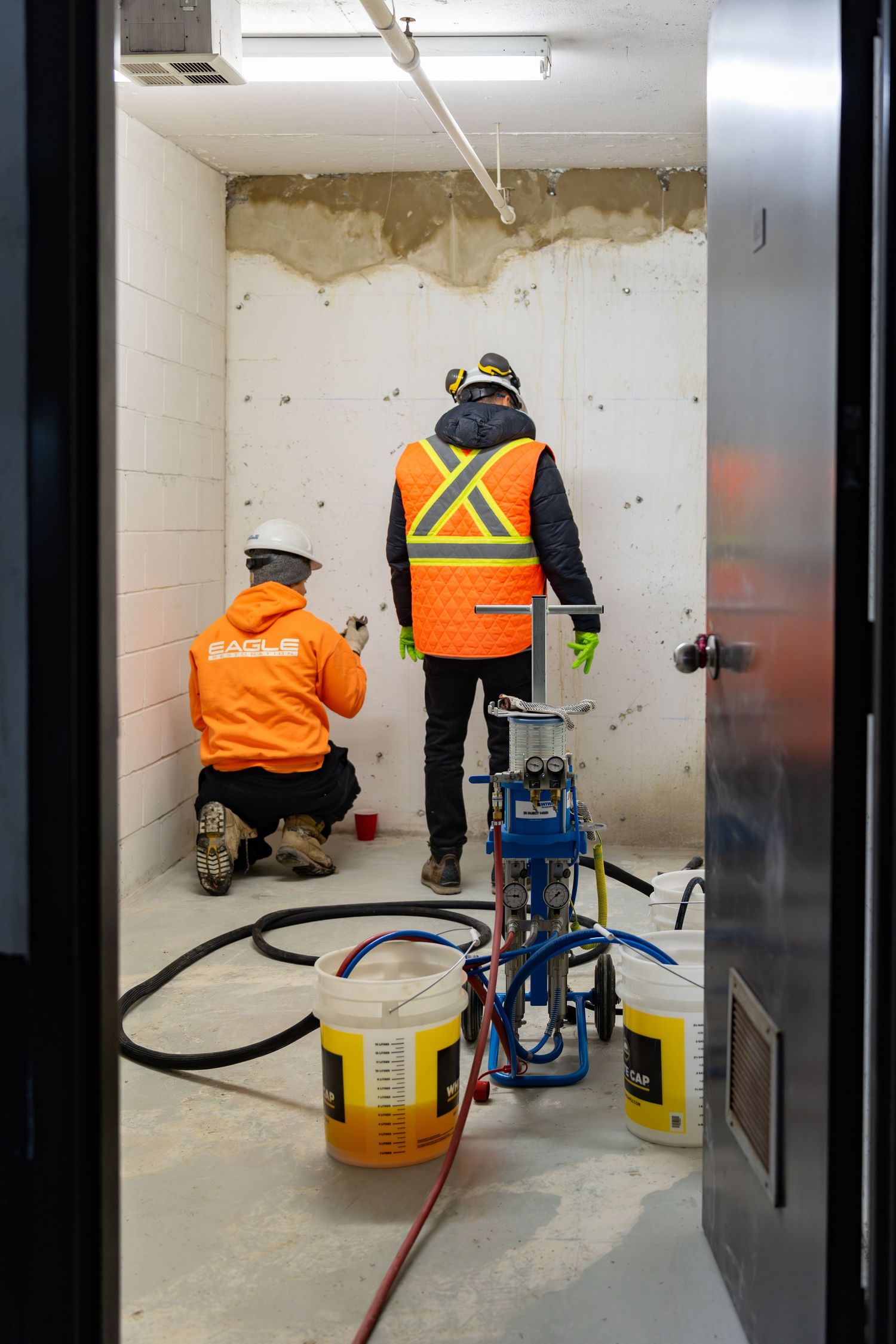 Construction workers in orange safety vests apply waterproofing sealant to a basement wall during renovation work.