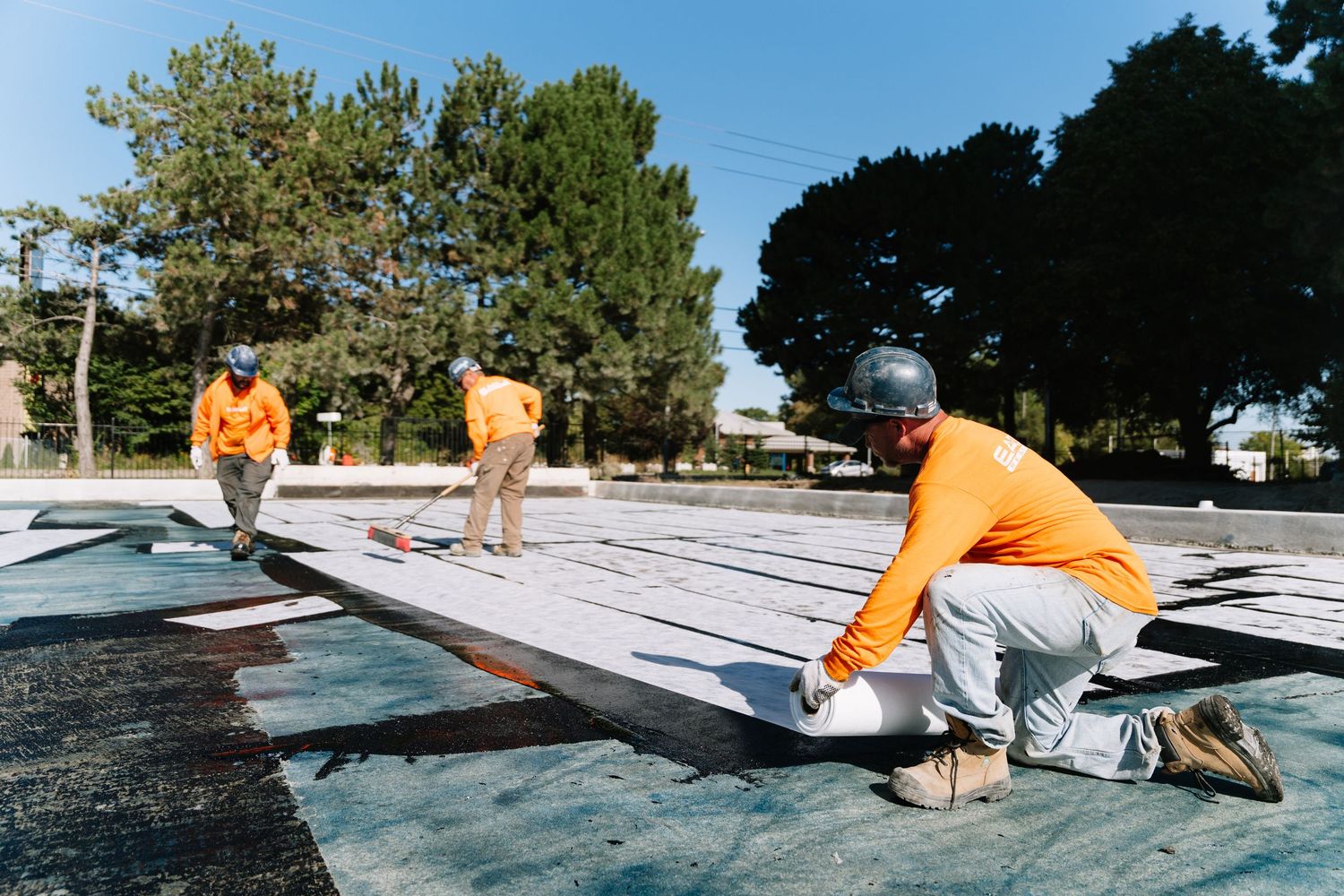 Construction workers in orange safety vests install white roofing material on a commercial building project.