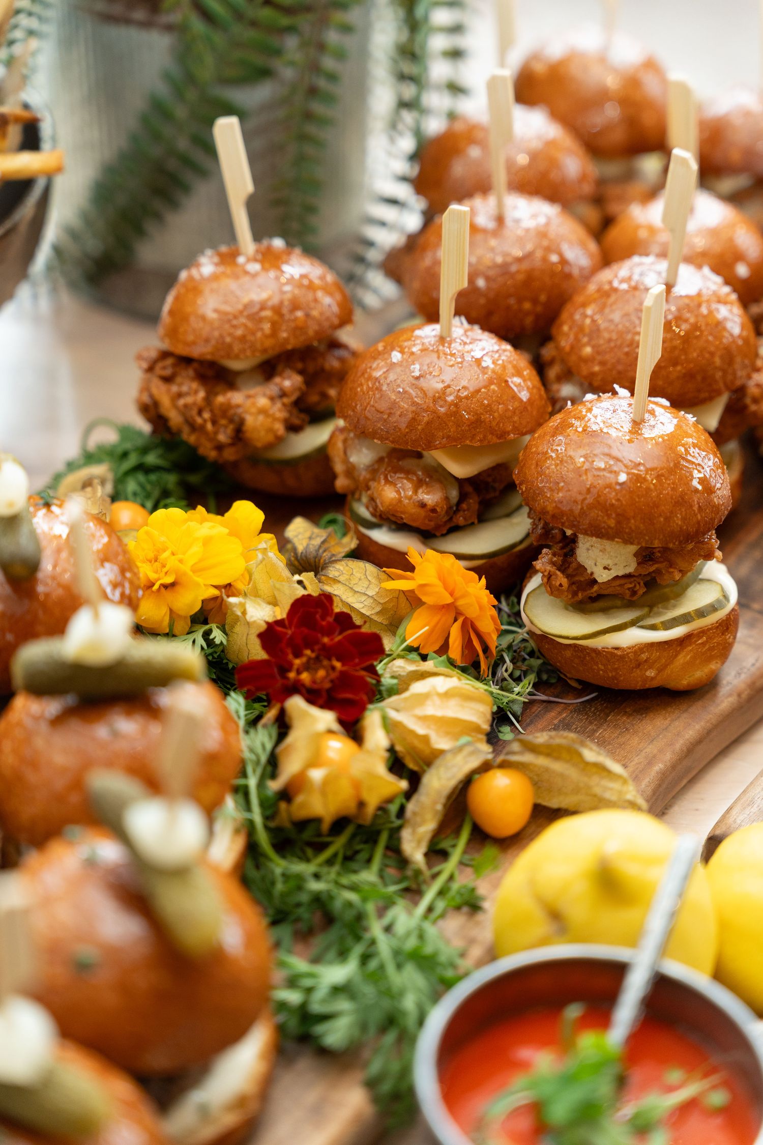Gourmet mini sliders with brioche buns served on a wooden board with colorful edible flowers and dipping sauce.