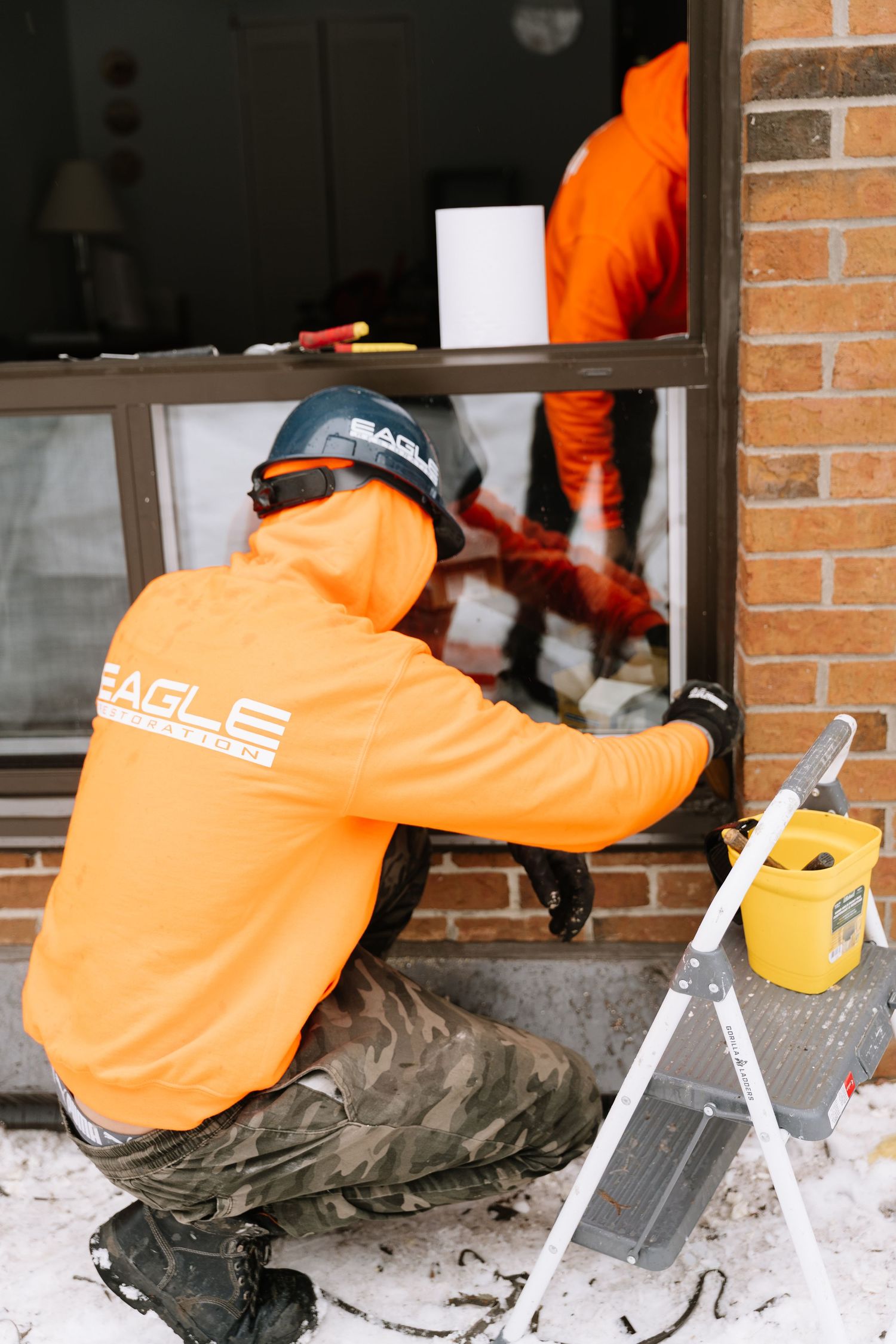 Construction workers in bright orange safety gear work on installing window frames in a brick building during winter.