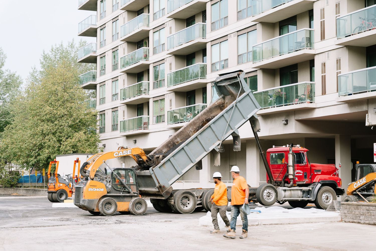 Construction vehicles and dump trucks lined up along an apartment building during renovation work.