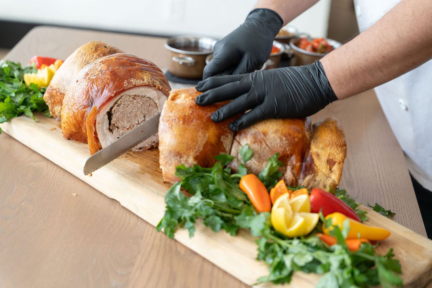 Hands wearing black gloves carve a golden roasted turkey garnished with fresh herbs and citrus on a wooden cutting board.