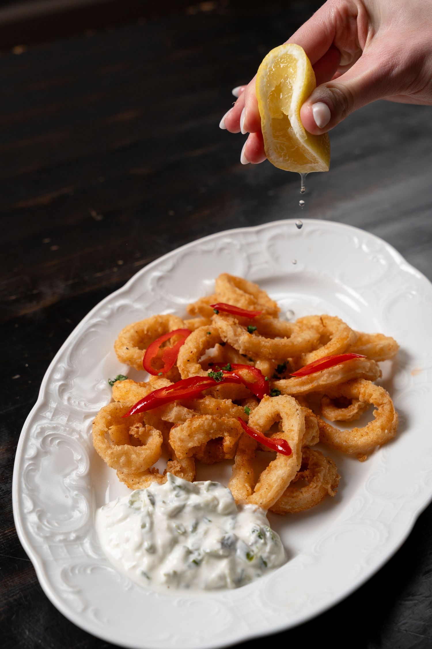 Hand squeezing lemon over crispy fried calamari rings with red chili and tzatziki dip on white plate.