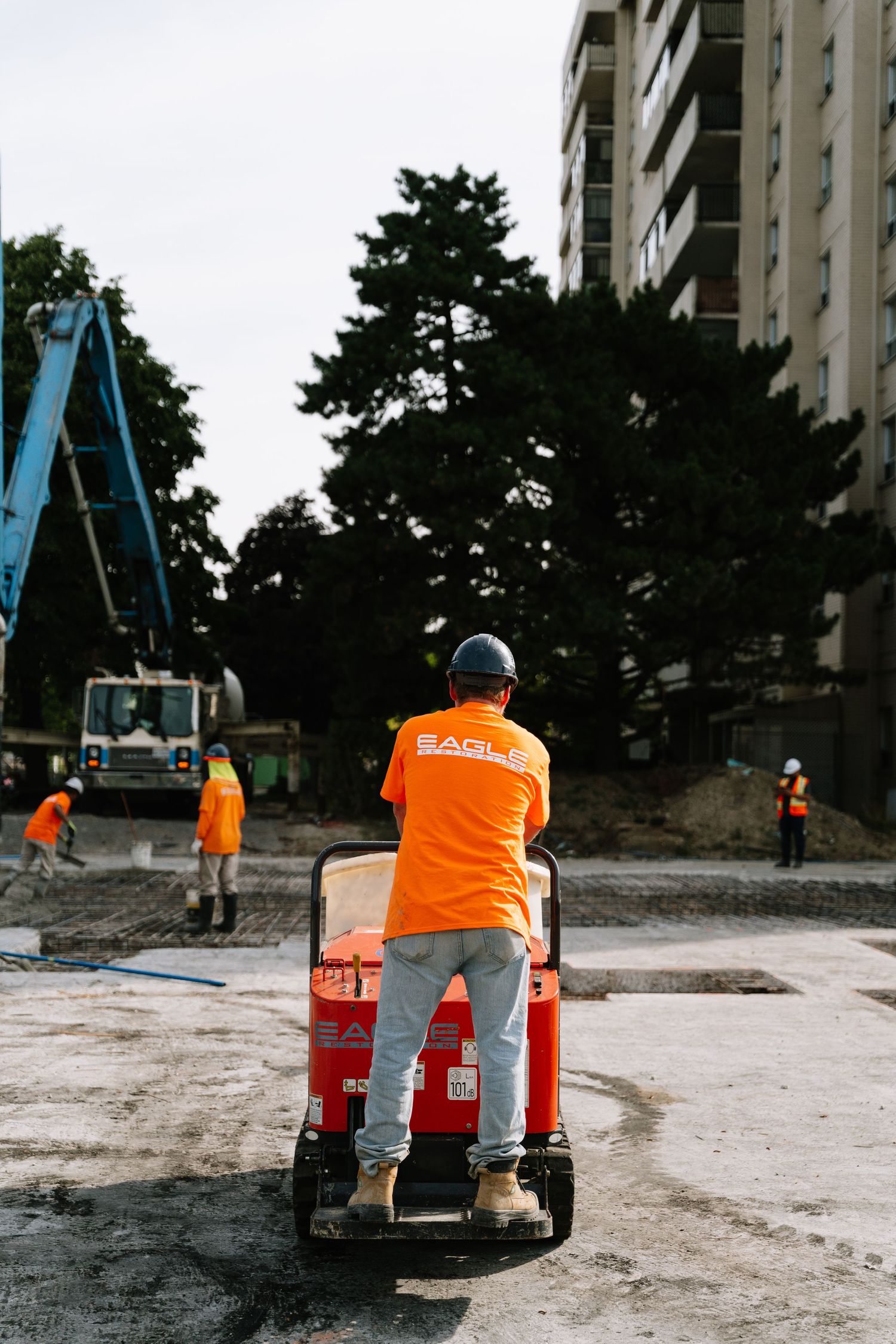 Construction worker in orange safety vest operates small equipment on dusty construction site near tall building.