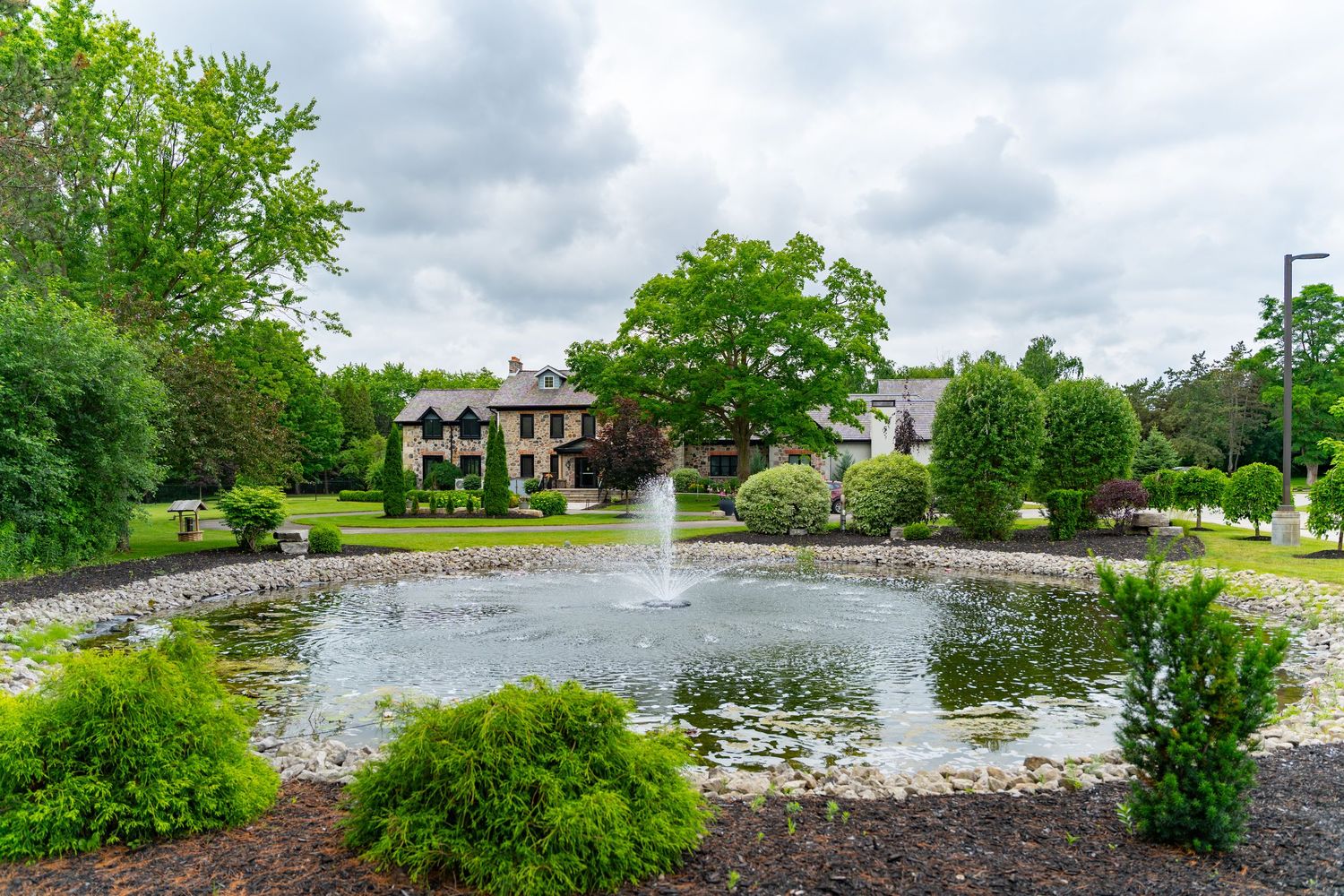 Lush green garden with a decorative pond fountain, manicured shrubs, and a stone manor house in the background.