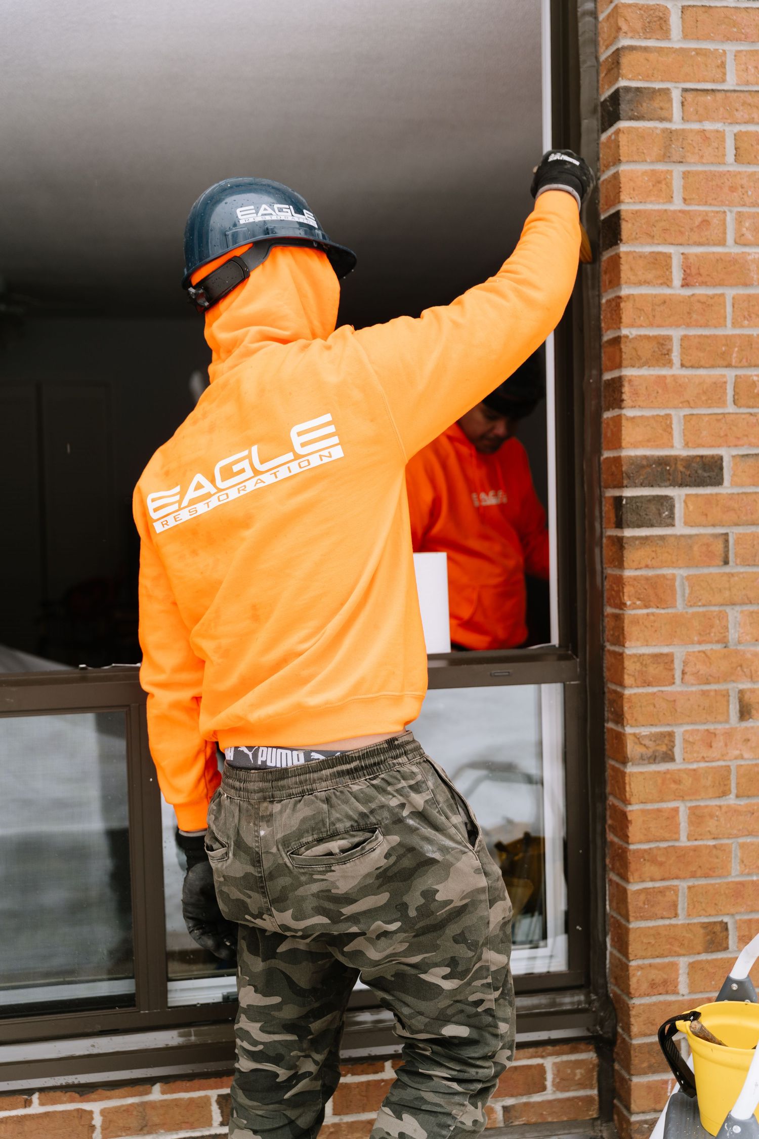 Worker in bright orange safety jacket and camo pants stands on ladder while performing window maintenance.