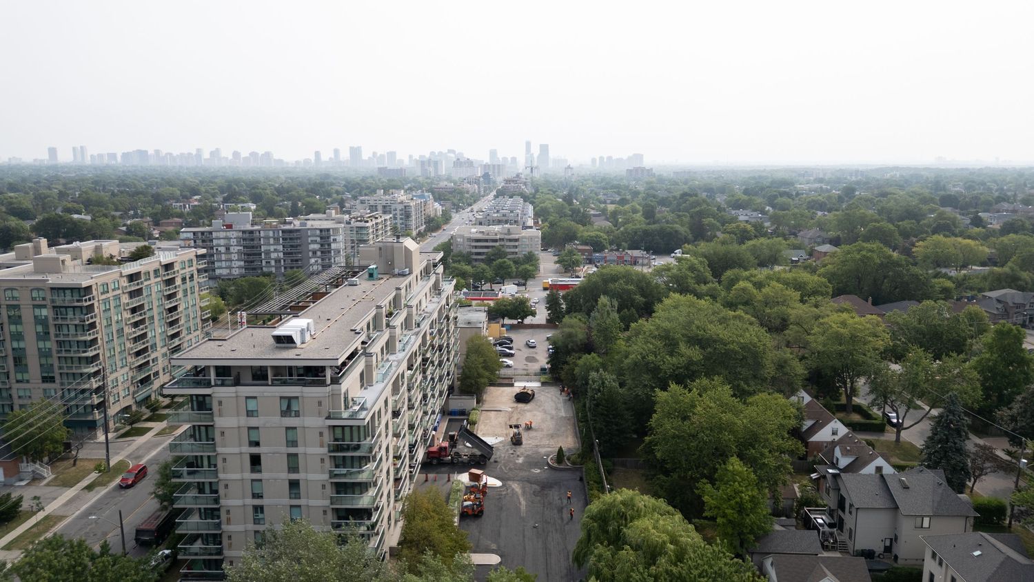 Aerial view of urban residential buildings and green tree canopy in a city neighborhood with downtown skyline visible.
