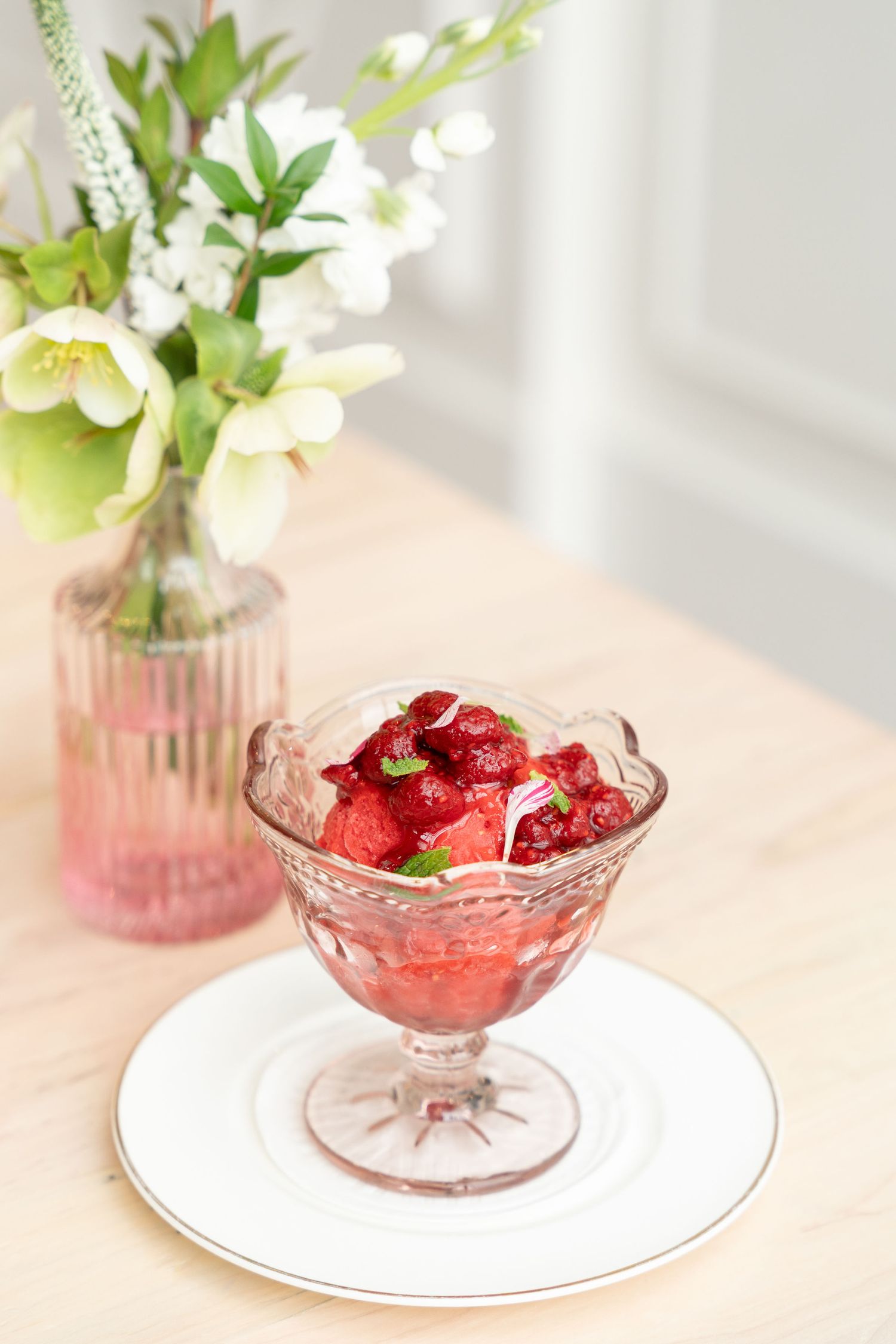 Crystal dessert bowl filled with red berries next to vase of white flowers on wooden table.