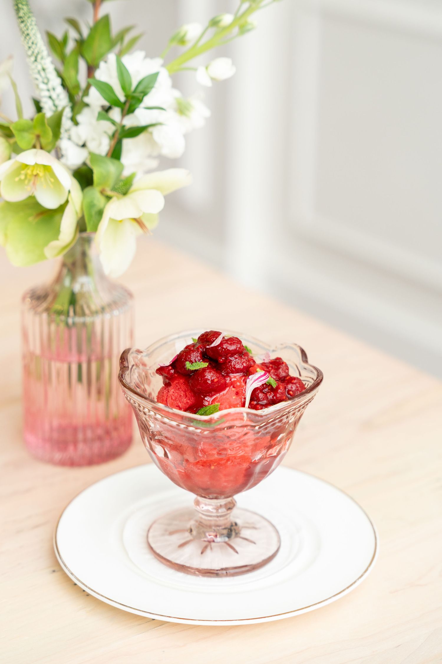 A crystal dessert bowl filled with fresh red cherries sits beside a pink vase with white flowers on a light surface.