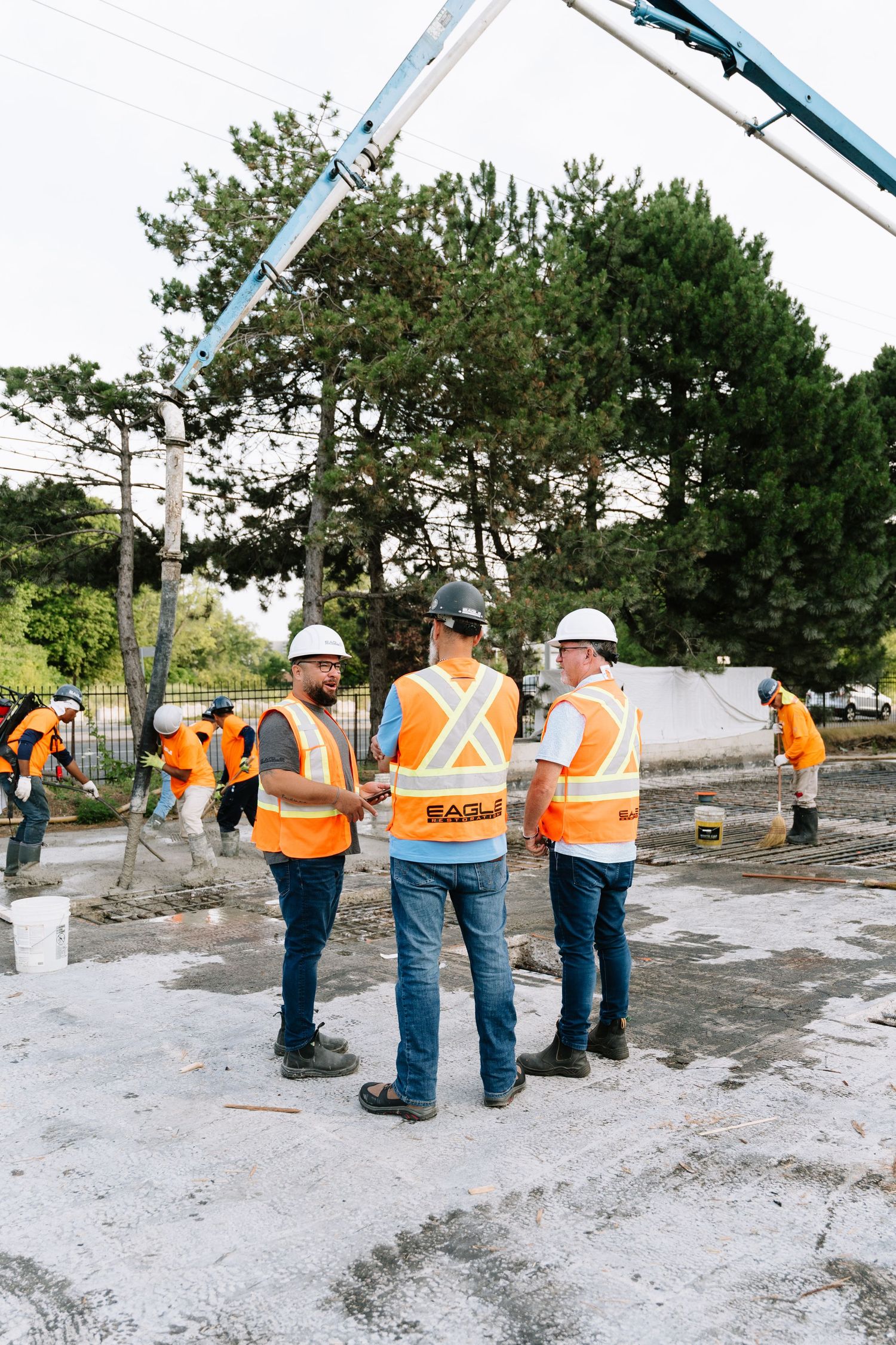 Construction workers in orange safety vests inspect and work on a concrete foundation at an outdoor worksite.