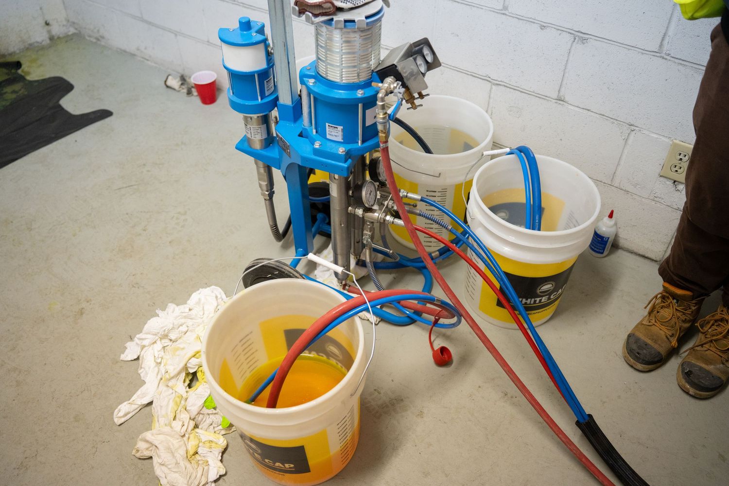 Reloading press setup with blue components and yellow buckets for ammunition reloading on concrete floor.