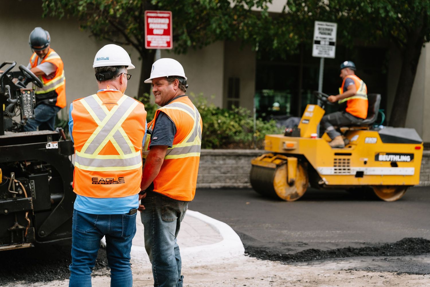 Road construction workers in safety vests supervise asphalt paving operations with steamroller in background.