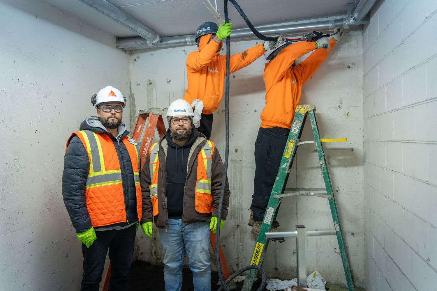 Construction workers in orange safety vests and hard hats inspect equipment in a concrete utility room.