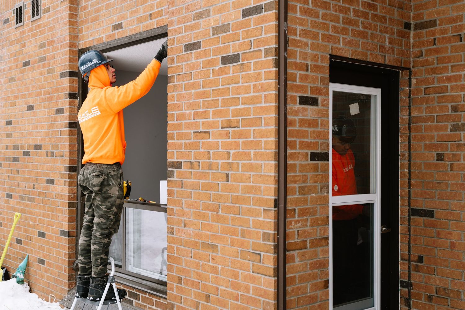 A maintenance worker in an orange safety vest installs a security camera on a brick building exterior.