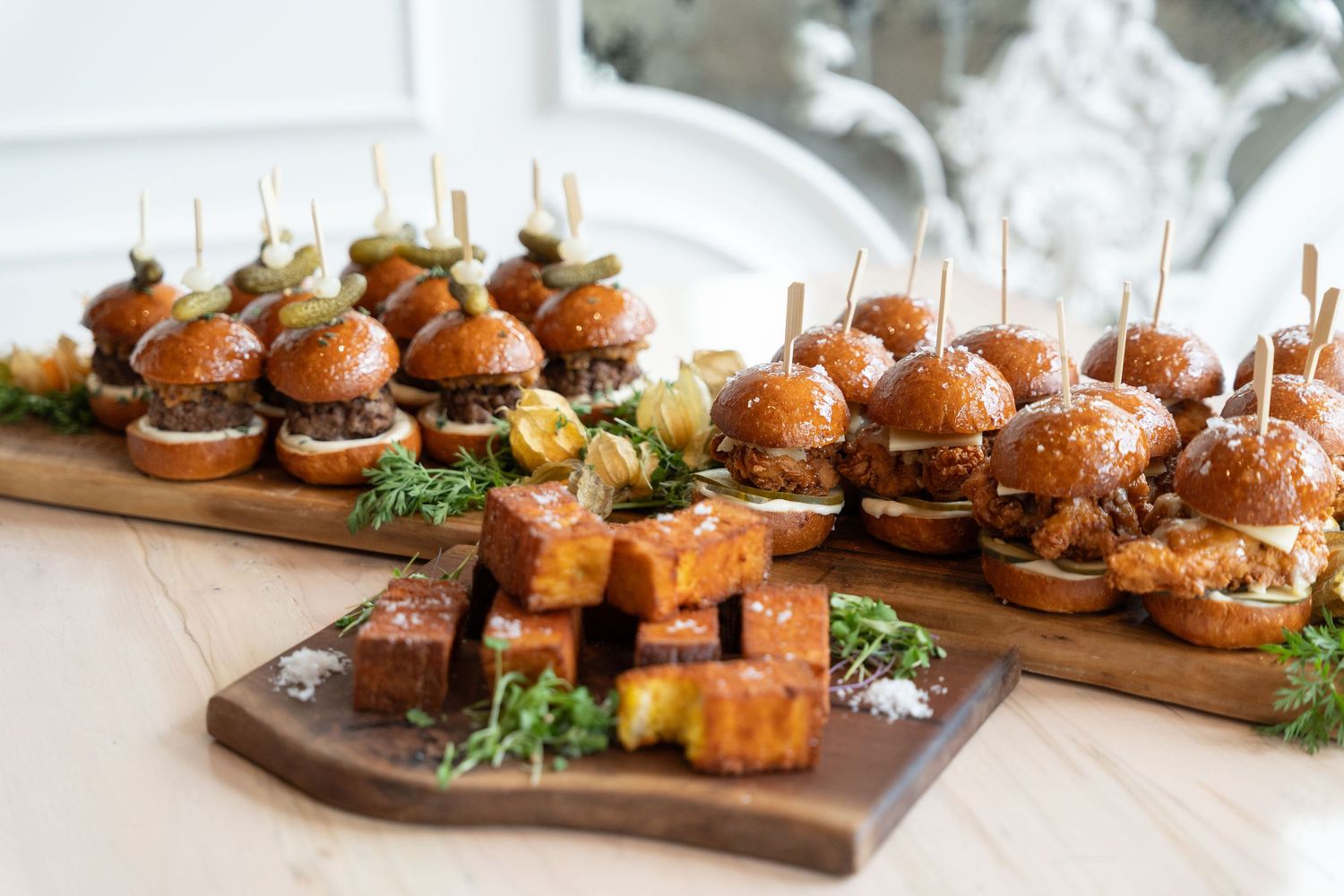 Wooden serving board with assorted gourmet sliders and appetizers arranged in a line for catering or events.