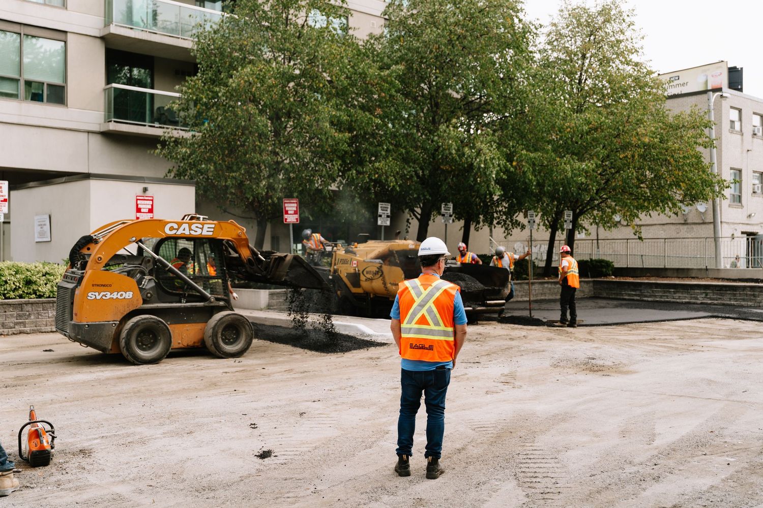 Construction worker in safety vest supervises equipment operation on urban street with trees and buildings.