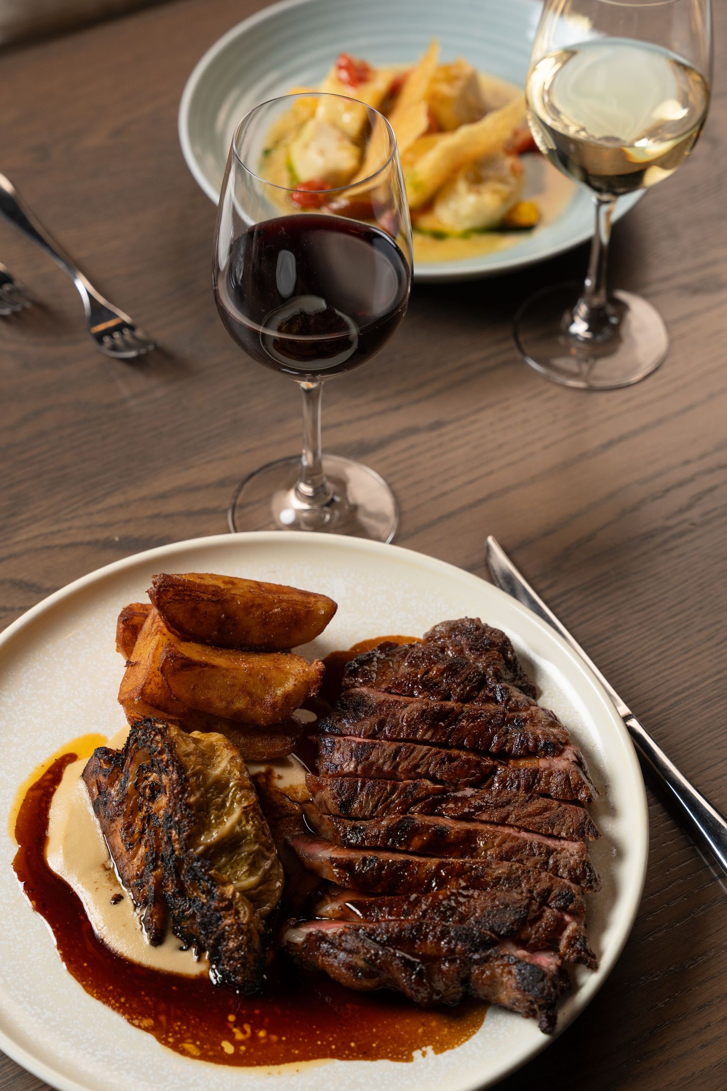 Grilled steak with roasted potatoes and sauce on a white plate, served with red and white wine glasses on a wooden table.