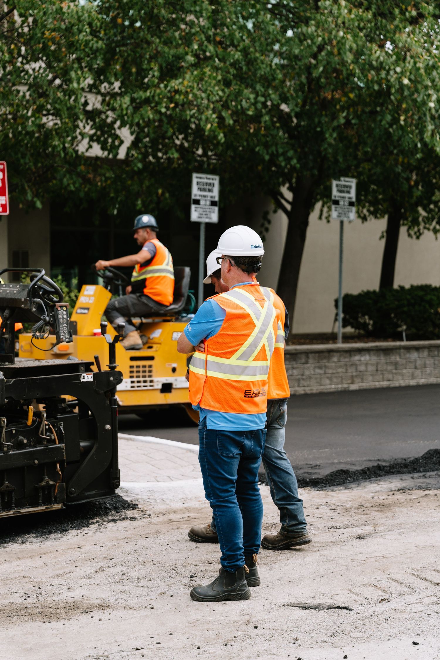Construction workers in orange safety vests operate paving equipment on a road resurfacing project.