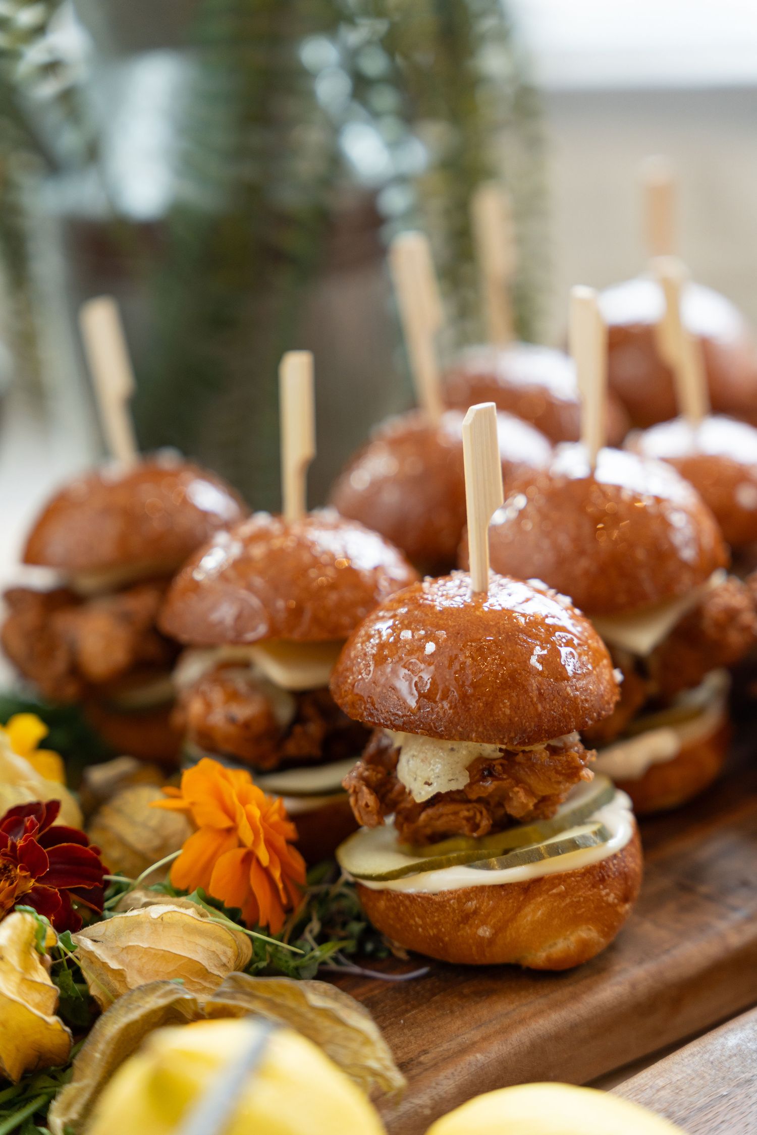 Row of gourmet mini sliders with fresh toppings arranged on a wooden serving board with colorful garnishes.
