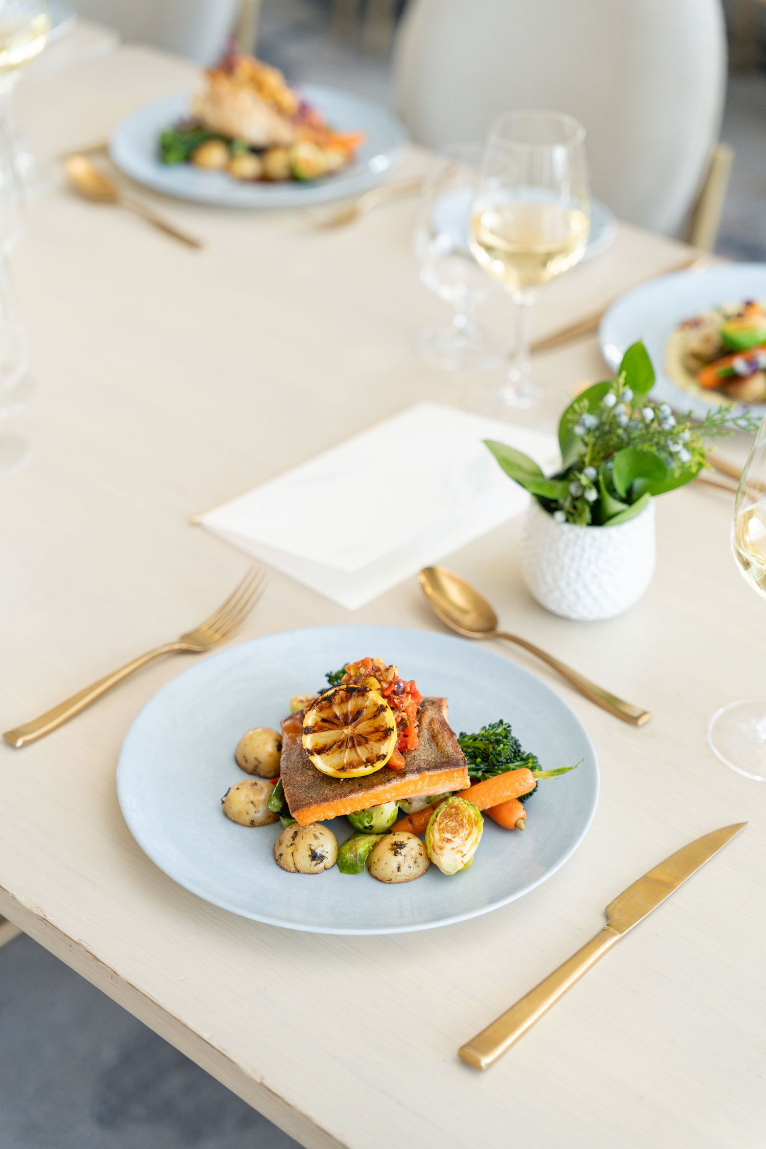 Elegant plated dish with grilled protein, roasted vegetables and gold cutlery on a white tablecloth with wine glass.