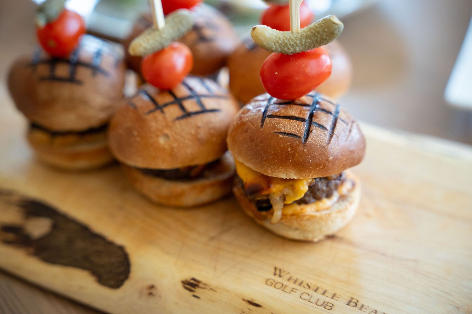 Mini gourmet sliders topped with cherry tomatoes and garnishes served on a wooden cutting board.