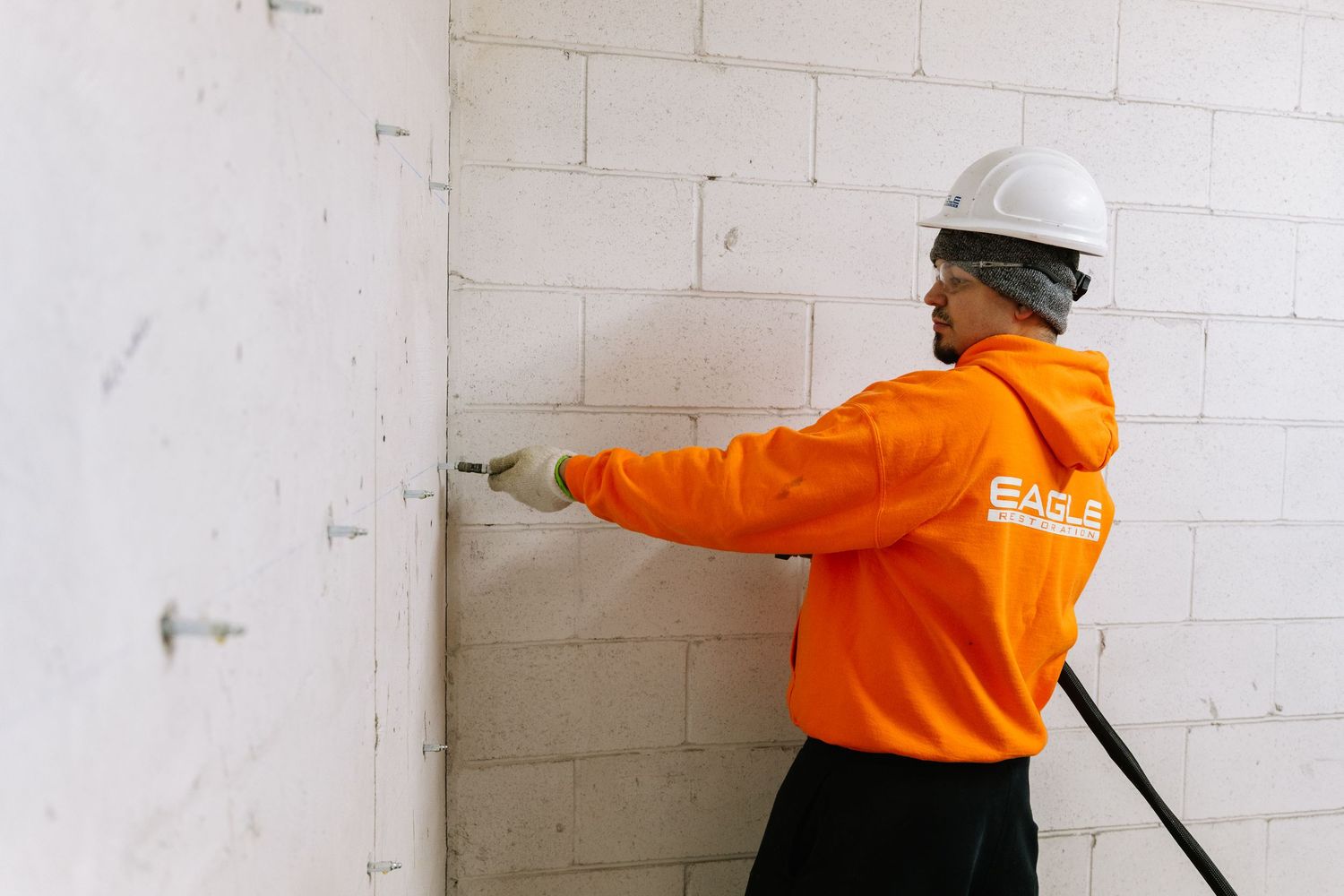 Construction worker in bright orange safety gear inspects white concrete block wall while wearing hard hat.