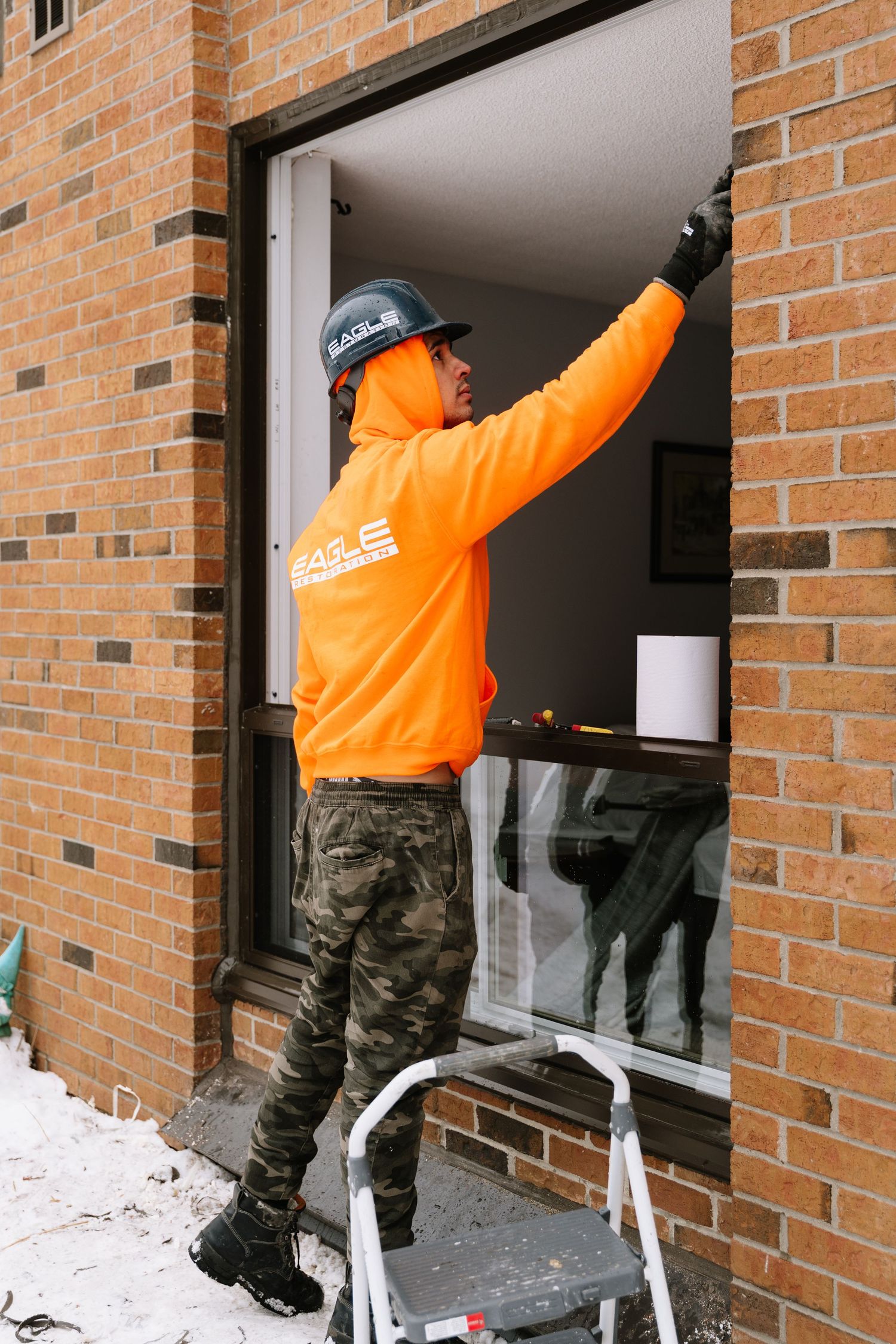 Window installer in orange safety gear works from ladder on brick building exterior.