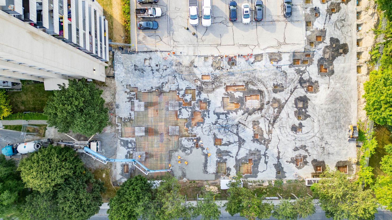 Aerial drone view of a parking lot with cars surrounded by buildings and trees on a sunny day.