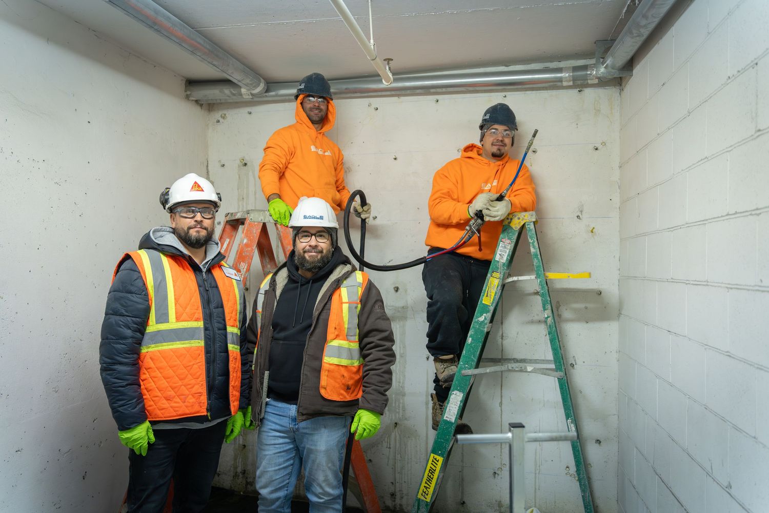 Construction workers in orange safety vests and hard hats collaborate on a project inside a concrete room.