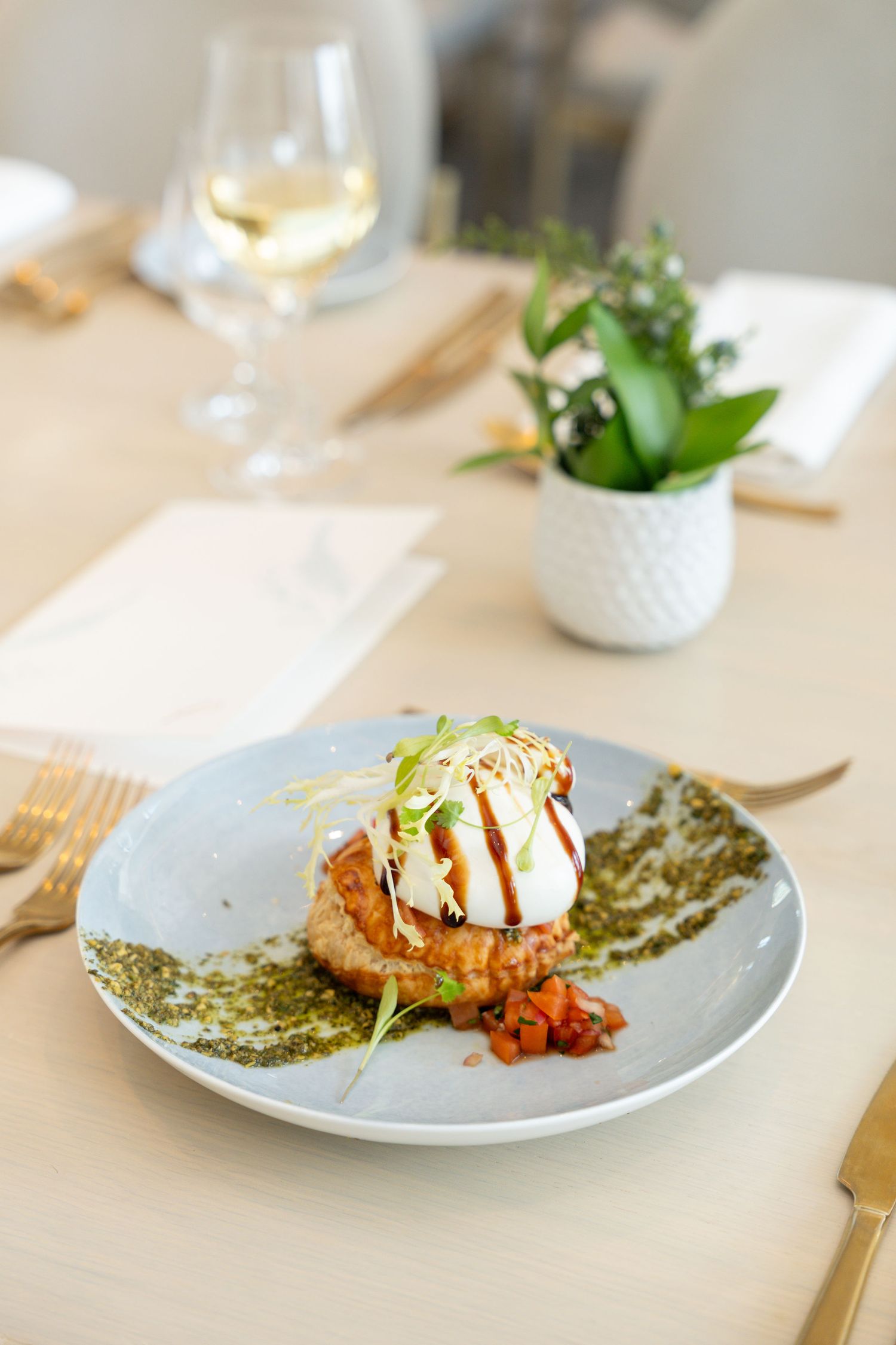 Elegant plated dish featuring a garnished protein with green sauce and microgreens alongside a glass of white wine.