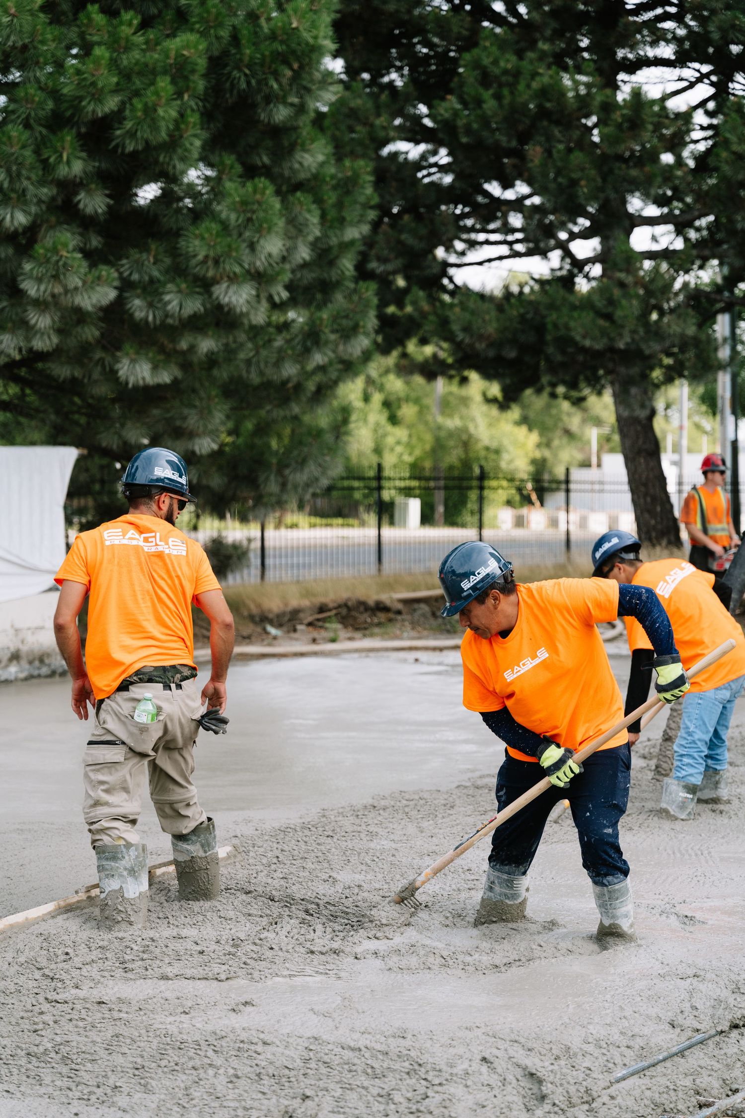 Workers in orange safety vests use tools to level and finish a fresh concrete surface at a construction site.