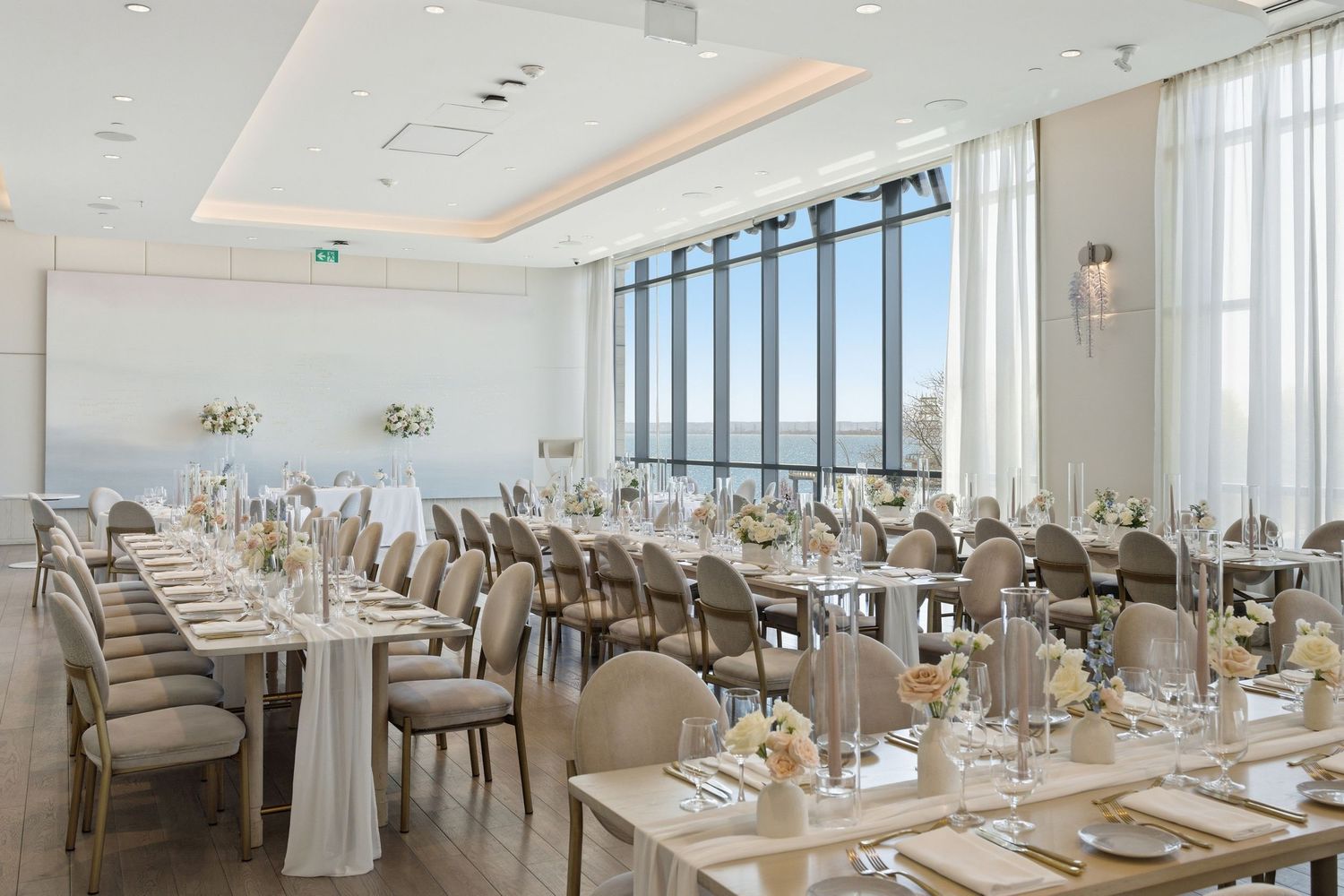 Elegant banquet hall with white chairs and tables set for dining, featuring panoramic windows and modern lighting fixtures.