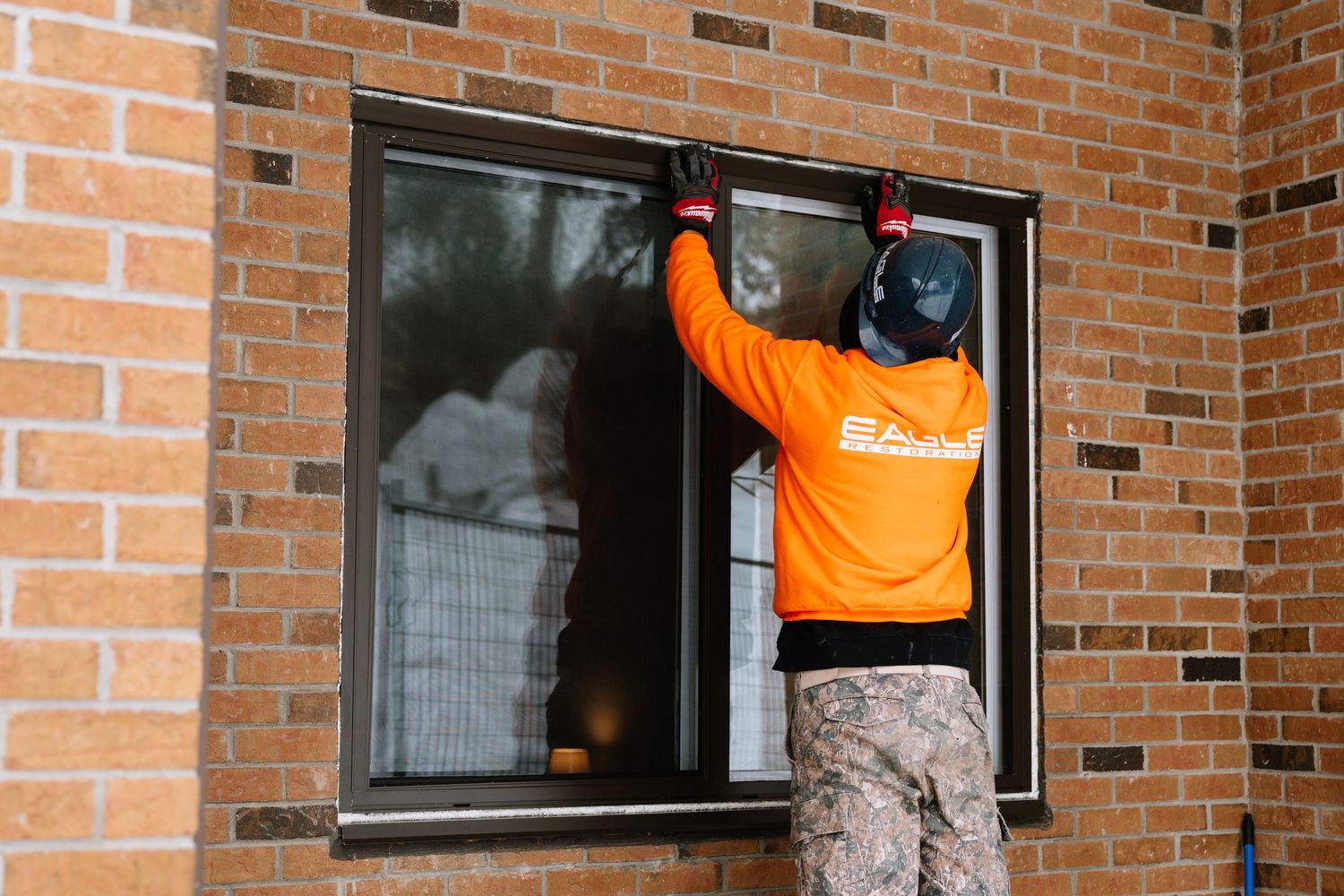 Worker in orange safety vest and camo pants stands on ladder while performing maintenance on brick building exterior.