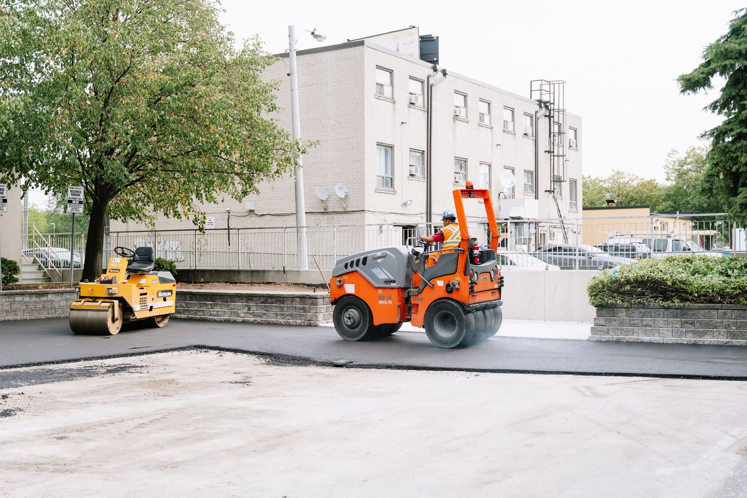 Orange compact construction vehicle operates on paved road near modern apartment building with trees.