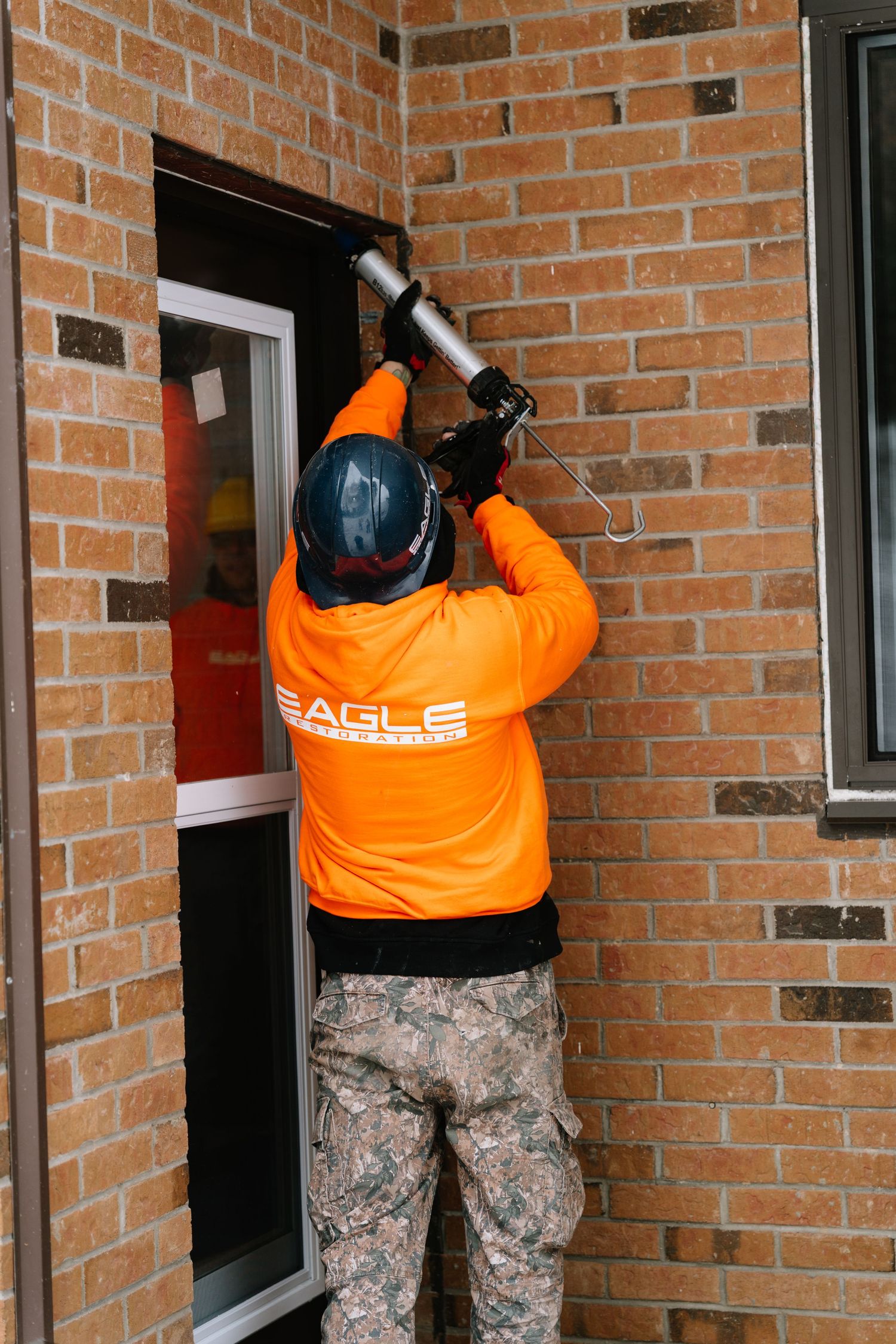 Maintenance crew member in orange safety gear works on multiple windows of brick building.