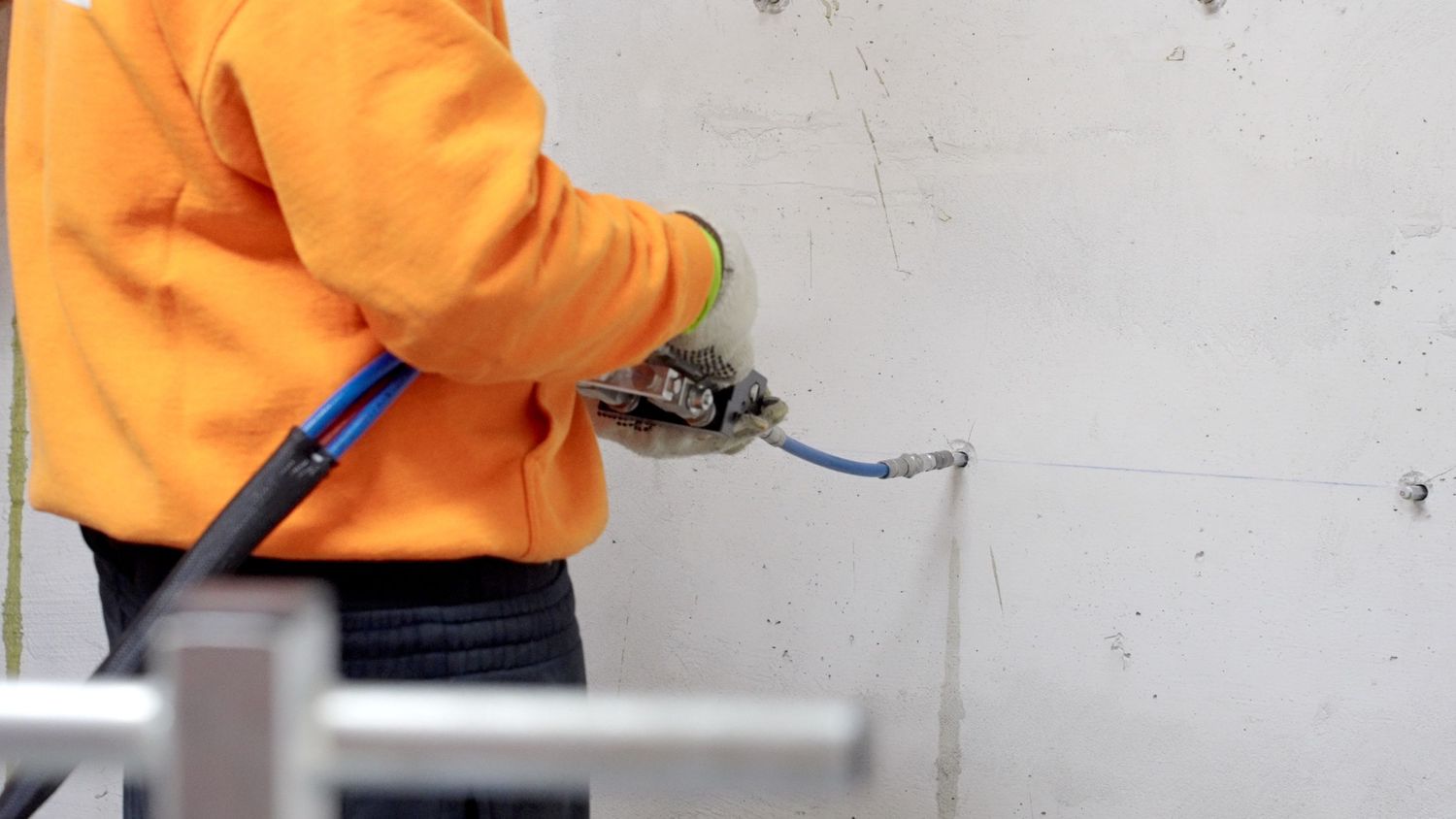 A worker in orange safety gear uses a power drill on a metal fixture mounted to a wall.