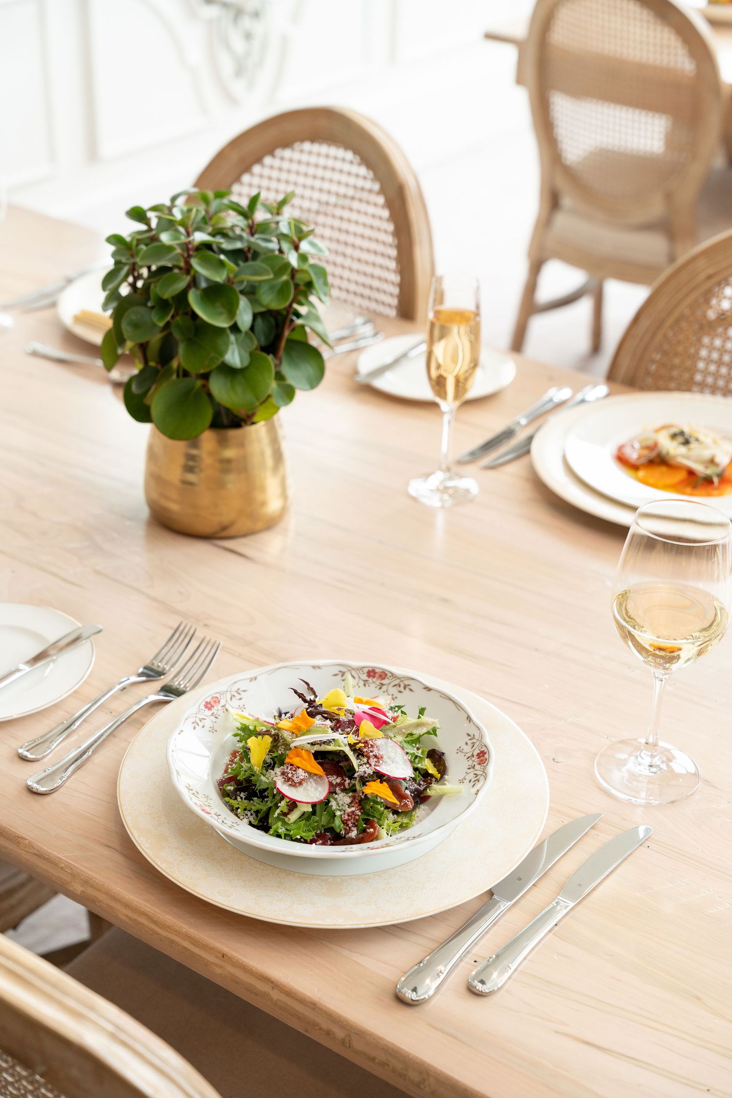 Fresh garden salad on white plate with brass plant holder and champagne glass on wooden dining table.
