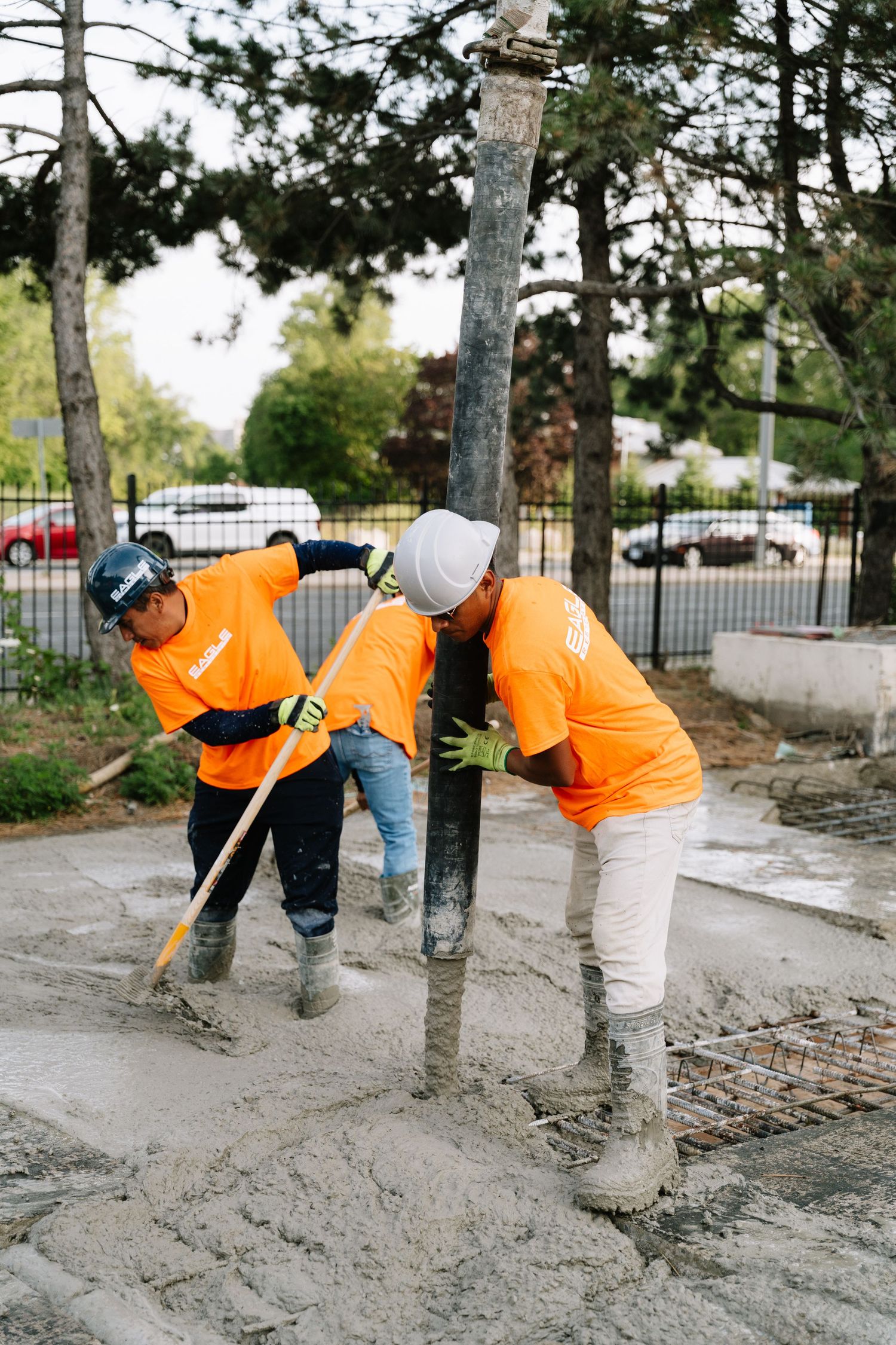 Workers in high-visibility orange shirts pour and level concrete on a construction site with trees in background.
