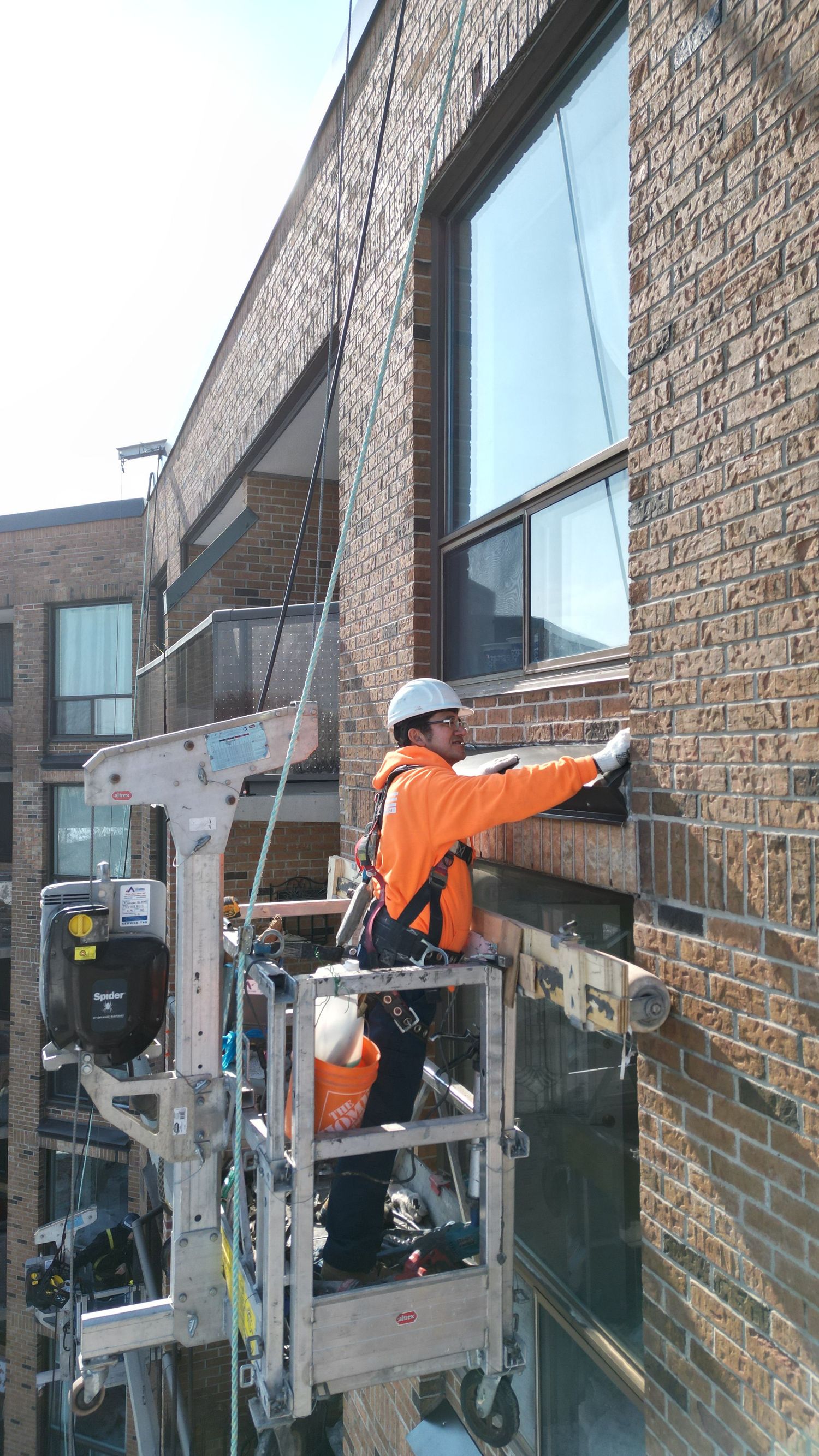 A maintenance worker in orange safety gear works from a suspended platform while cleaning windows on a brick building.