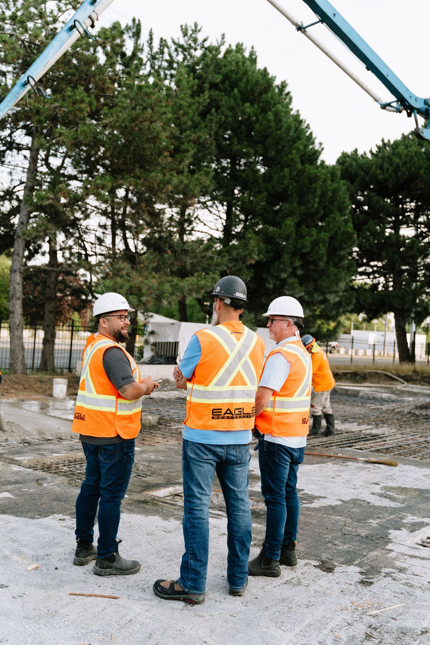 Construction workers in orange safety vests and hard hats stand in line on a concrete surface near pine trees.