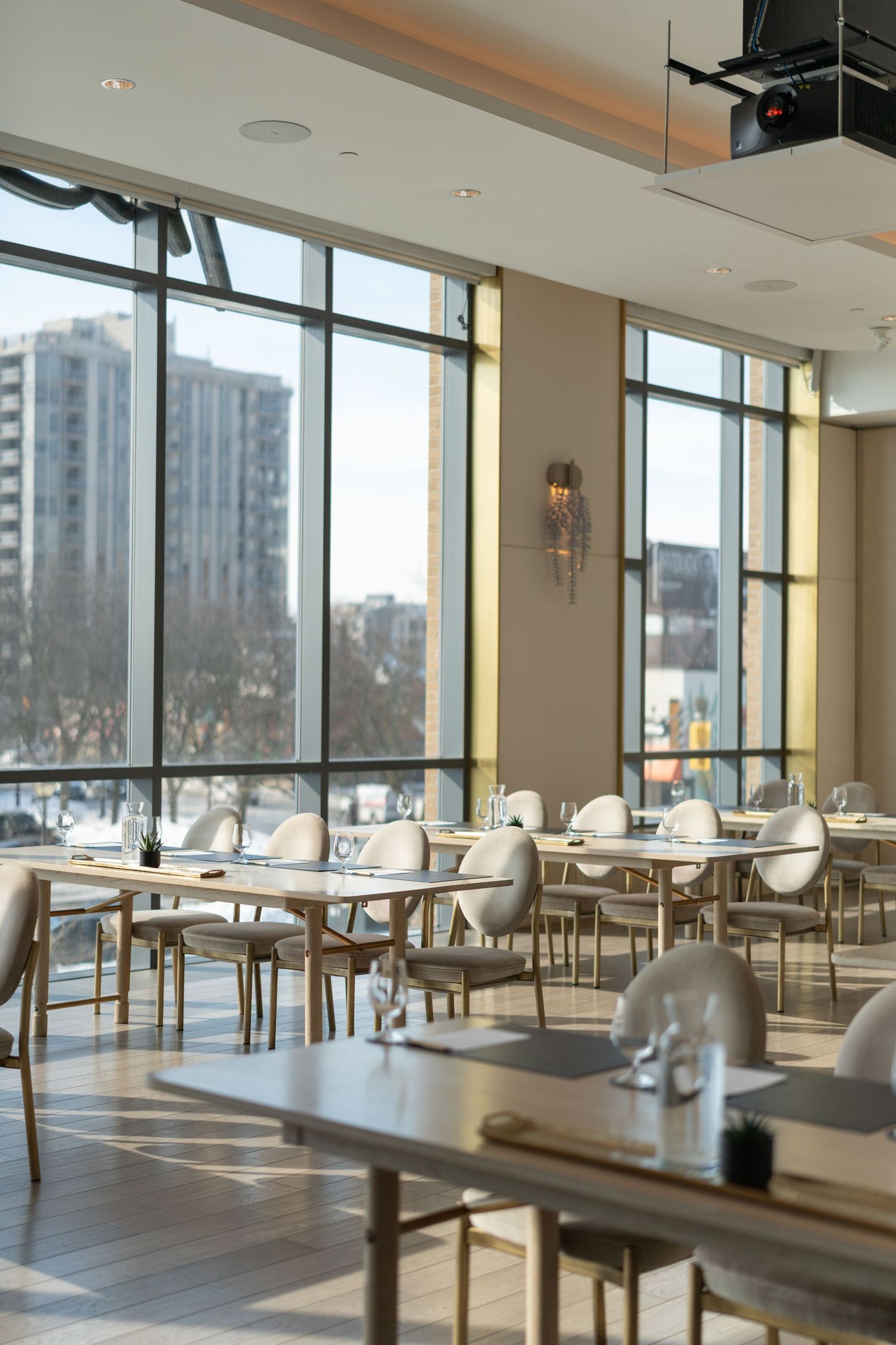Modern restaurant interior with floor-to-ceiling windows, white tables and chairs, and city views in natural daylight.